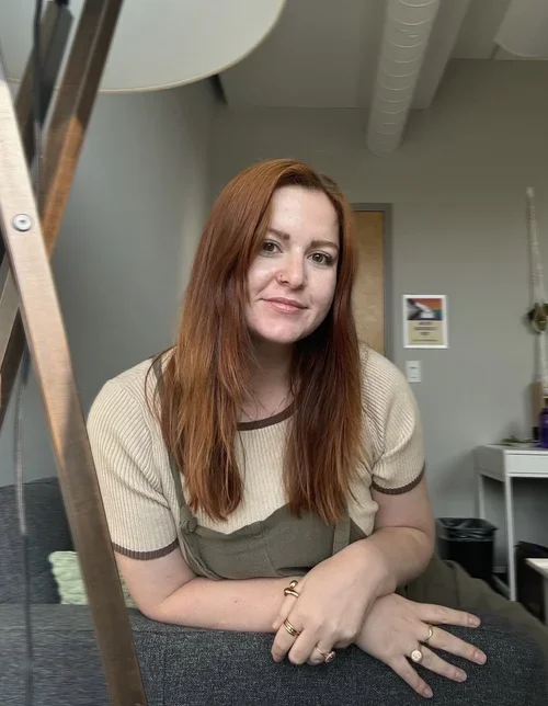 A young woman with long red hair sitting indoors, smiling at the camera.