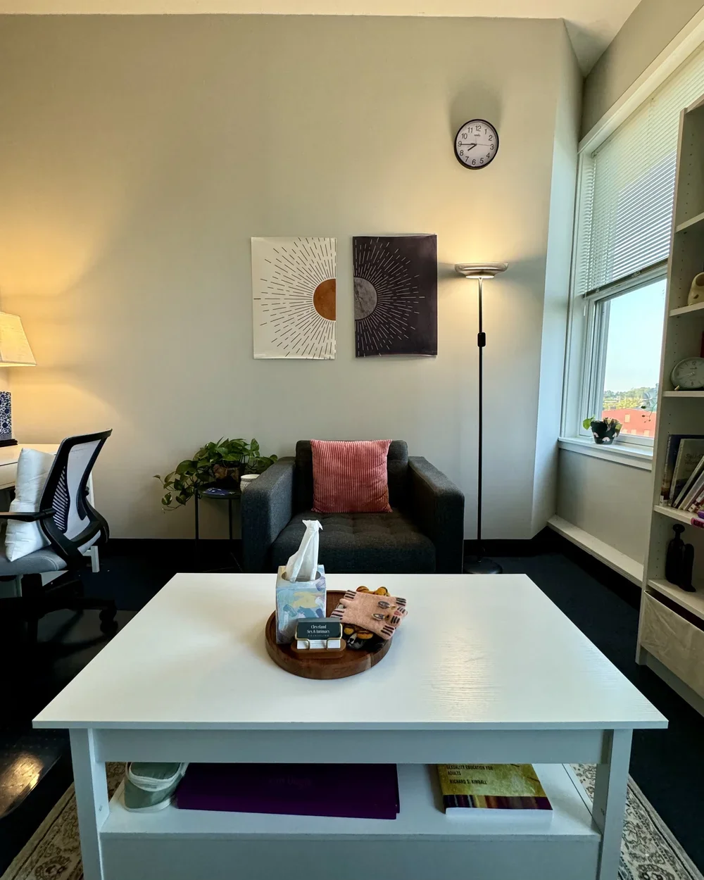 A cozy office with a white table, gray armchair, and black office chair. Artwork depicting sun and moon hangs on the wall. A floor lamp and a window near the right wall provide natural light. The table has tissues, a tray with small items, and a book