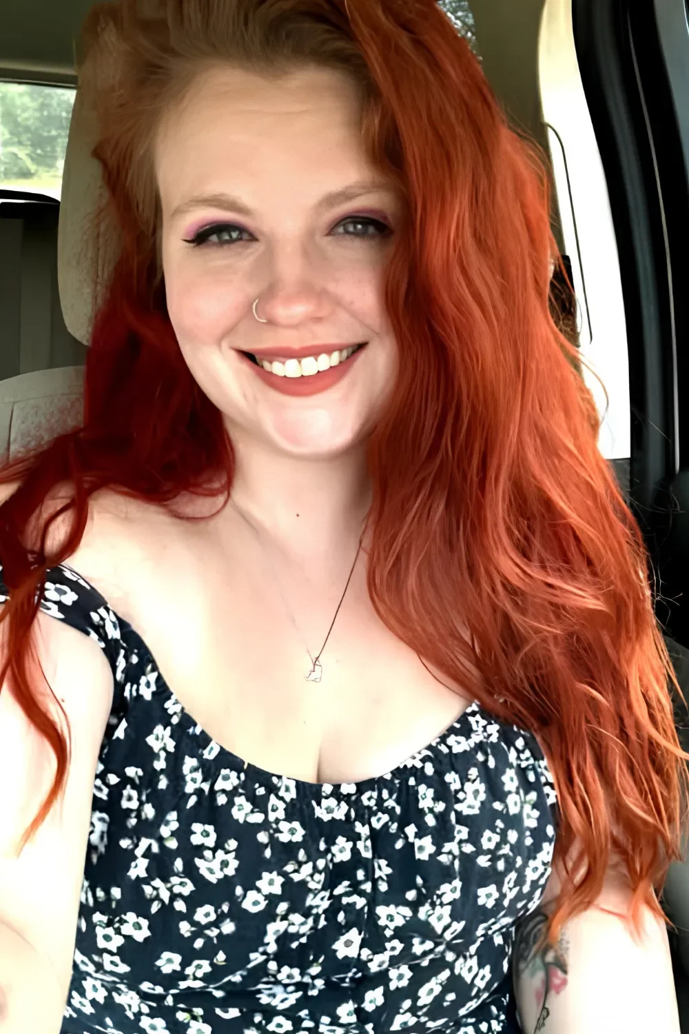 A woman with long, wavy red hair smiling inside a car, wearing a black and white floral top, with a necklace and a nose ring.