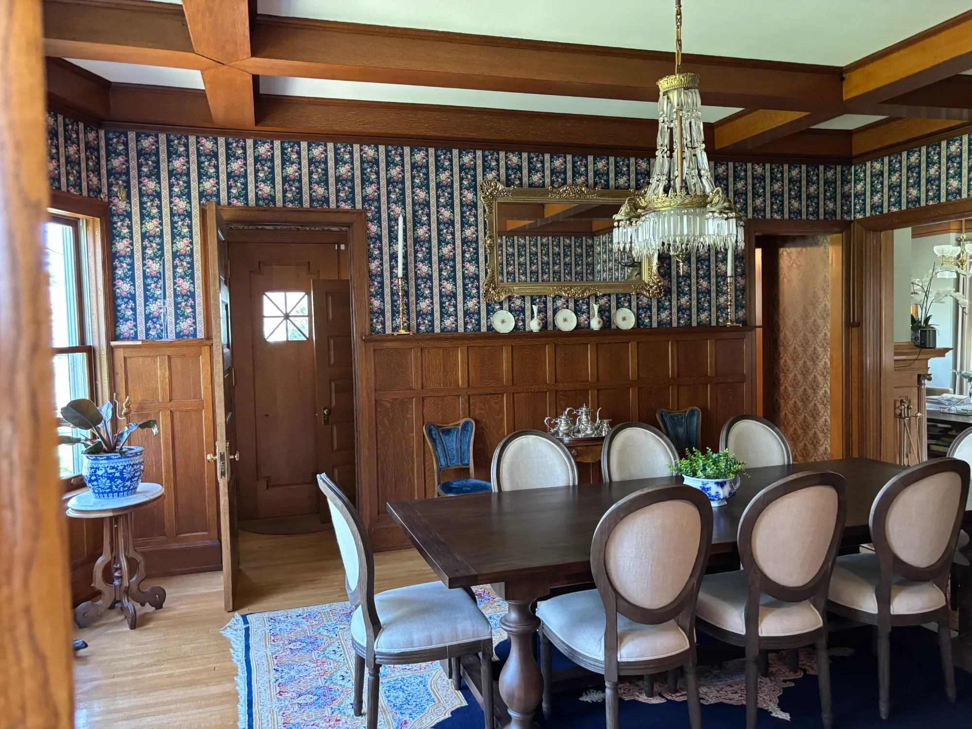 A dining room with floral wallpaper, a chandelier, wooden accents, and a table with beige chairs. A blue and white plant pot is on a small side table near a window.