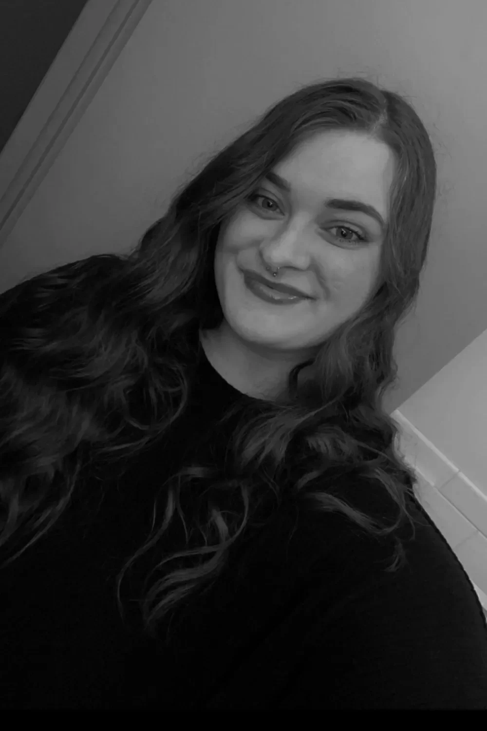 A Black and White photo of a woman with long, curly hair, smiling, wearing a septum piercing, and a dark top, standing indoors near a door.