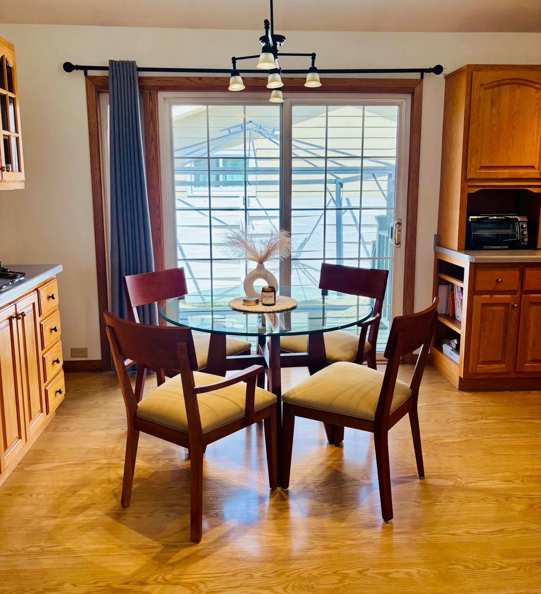 A dining area with a round glass table, wine-colored chairs with beige cushions, a window with wood trim, and a view of an outdoor patio with a large umbrella frame. Wooden cabinetry is on the right side, and a chandelier hangs above the table, with sunlight entering through the window.