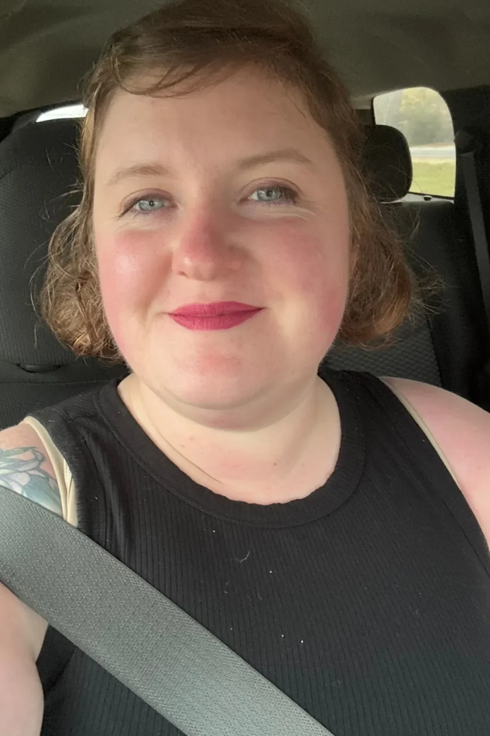 A woman with short, wavy red hair, wearing a black sleeveless top, smiling, sitting in a car with a seatbelt on.