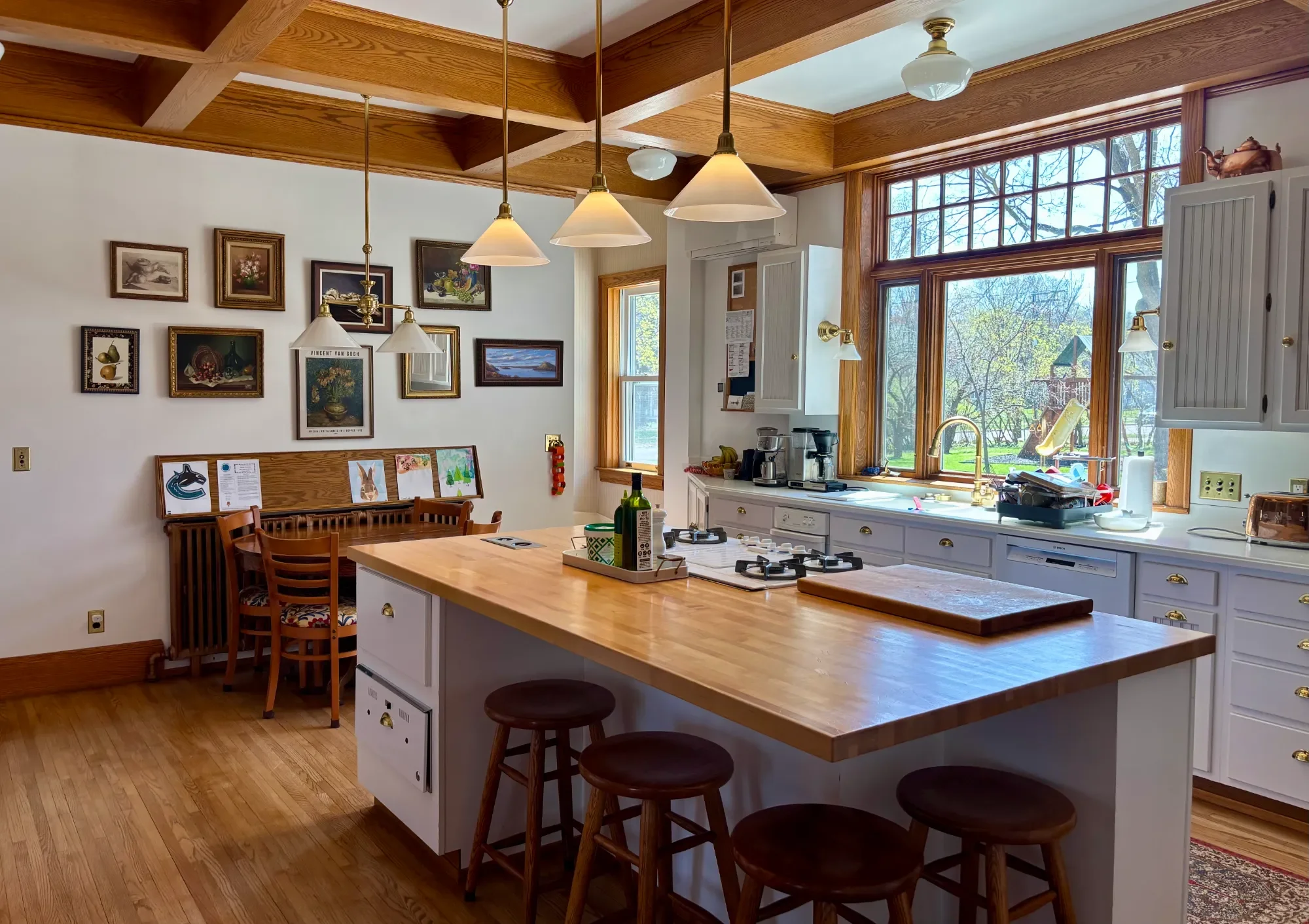 Kitchen with large wooden-framed windows, white cabinetry, a wooden center island, and pendant lights hanging from a wood coffered ceiling. The room has hardwood floors and decorative framed artwork on the wall.