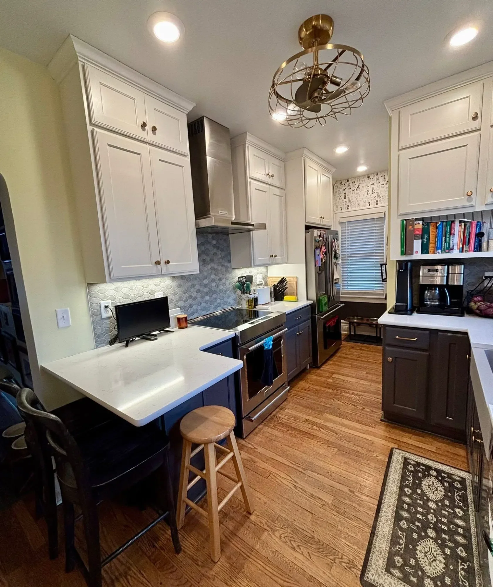Kitchen with white cabinets, stainless steel stove, gray backsplash, black and gray lower cabinets, small computer monitor on white island, wooden stools, floral patterned rug, bookshelves with cookbooks, and a window with white blinds.