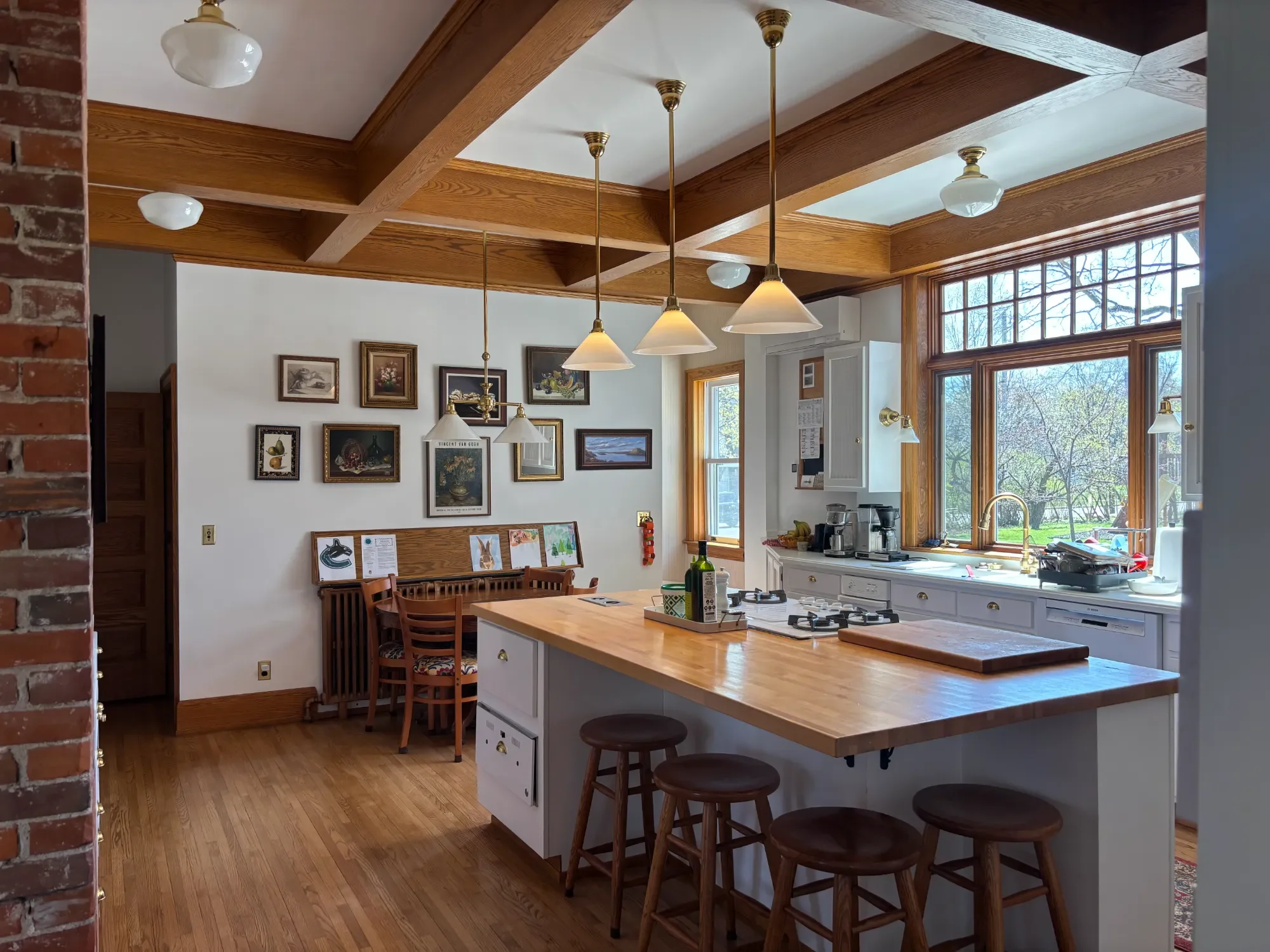Kitchen with a wooden coffered ceiling, white cabinets, large window with wooden trim, hanging pendant lights, and a wooden dining table with chairs.