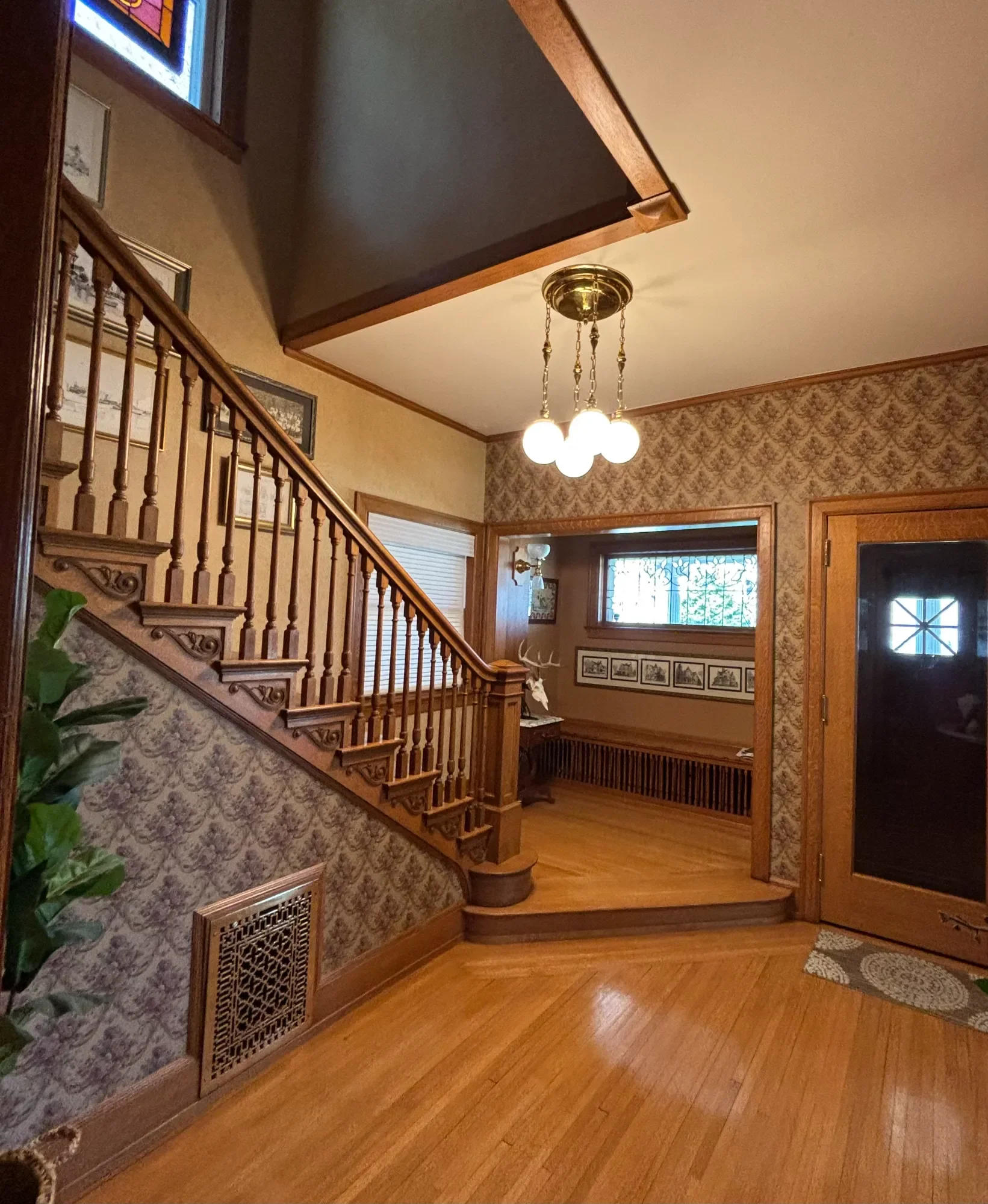 Interior view of a home with hardwood floors, wood staircase with spindles, a decorative wall with framed artwork, a window with blinds, and a light fixture with multiple globes hanging from the ceiling.