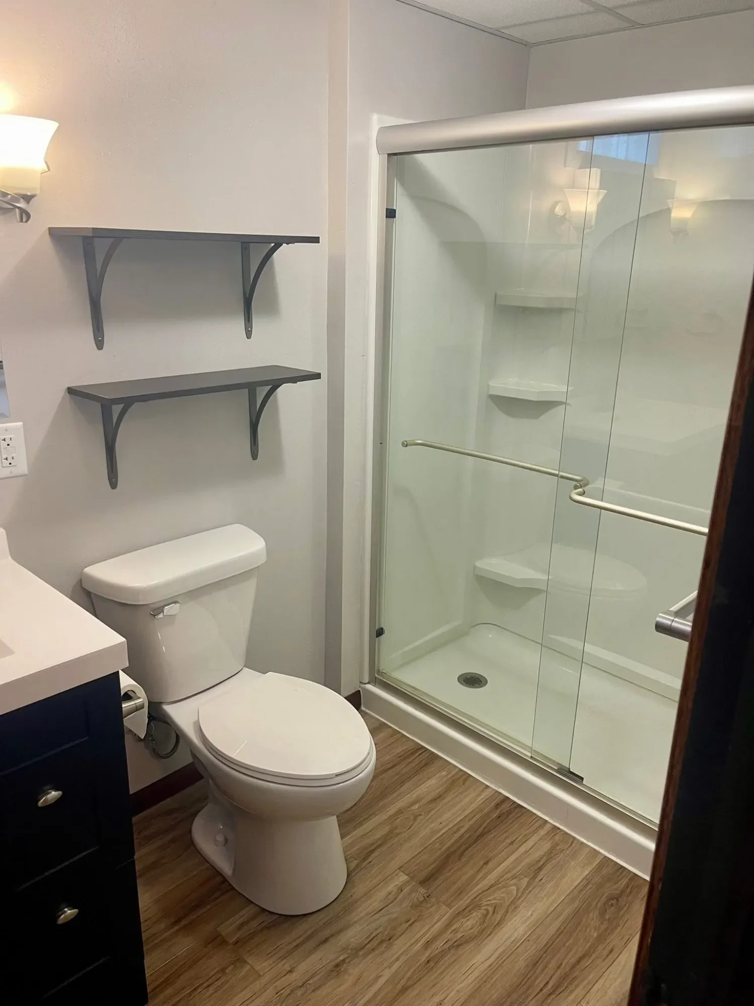 A bathroom with a toilet, black vanity with drawer, two wall-mounted shelves, and a glass shower with shelves and a sliding door.