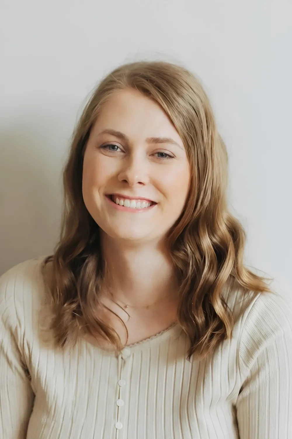 A young woman with shoulder-length wavy light brown hair, smiling and looking at the camera, wearing a cream-colored ribbed top with buttons, standing against a plain light background.