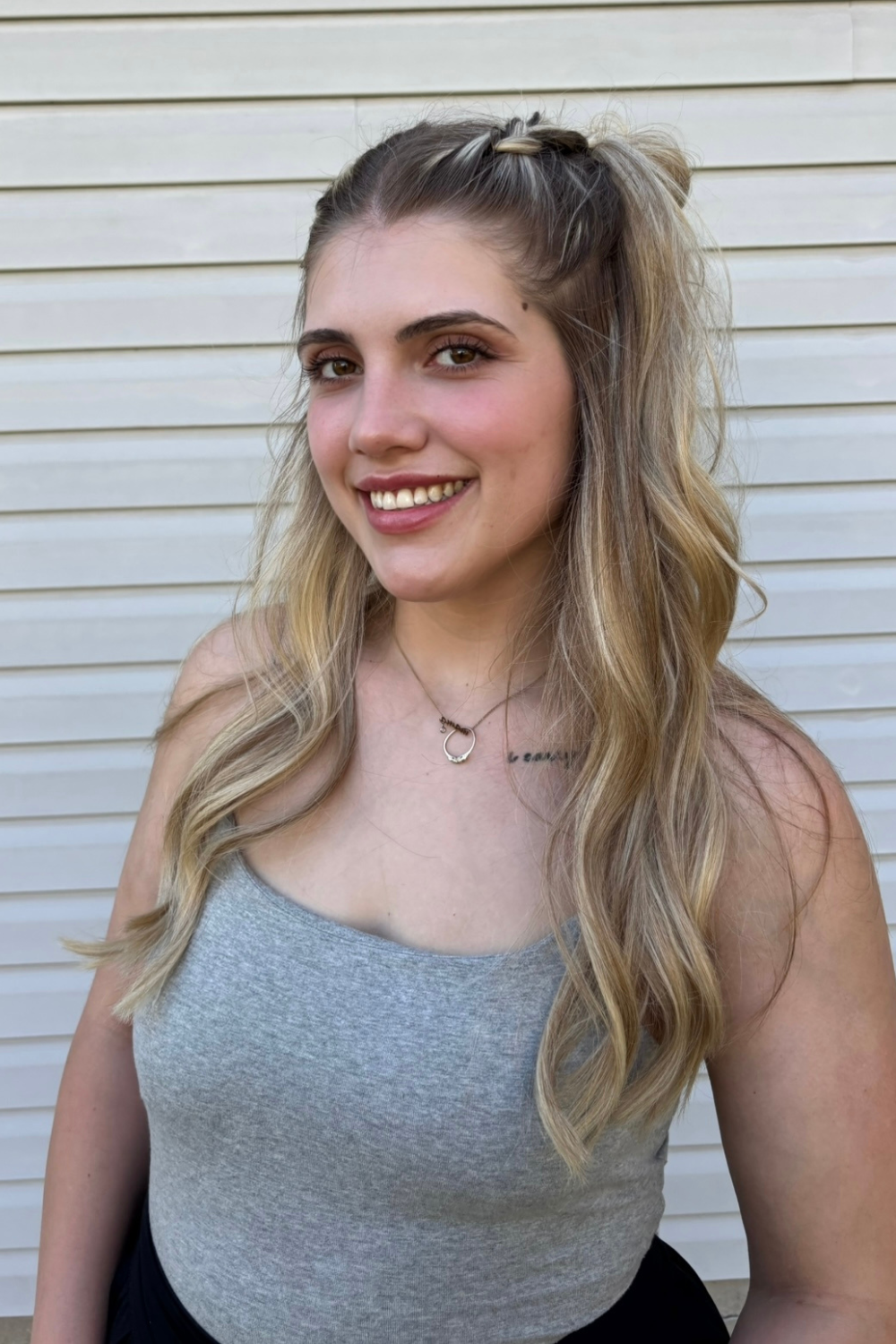A young woman with long, wavy blonde hair, smiling, wearing a gray sleeveless top and a necklace, standing in front of a white horizontal siding wall.
