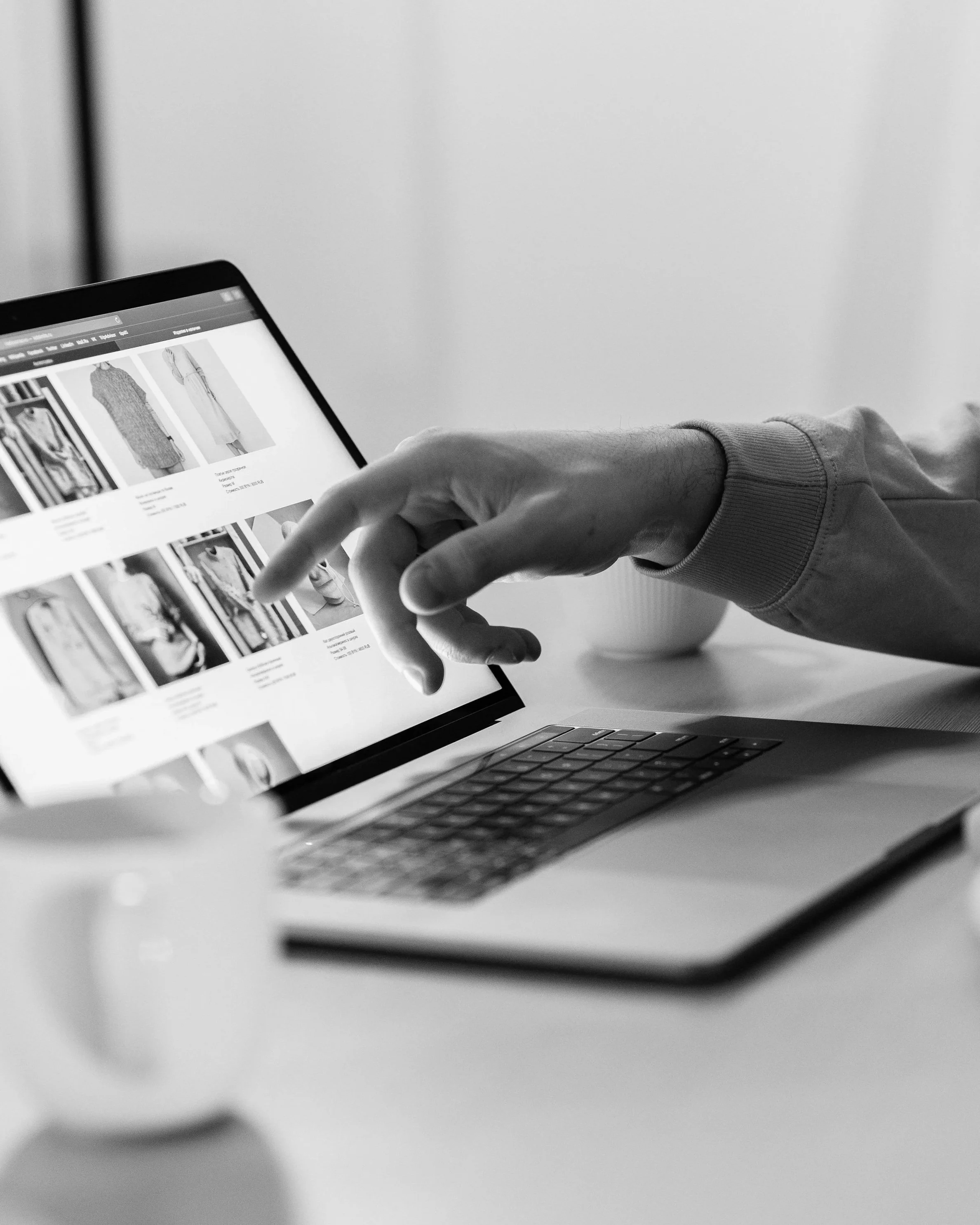 Black and white photo of a person using a laptop to browse a clothing website, with their hand and finger pointing at the screen.