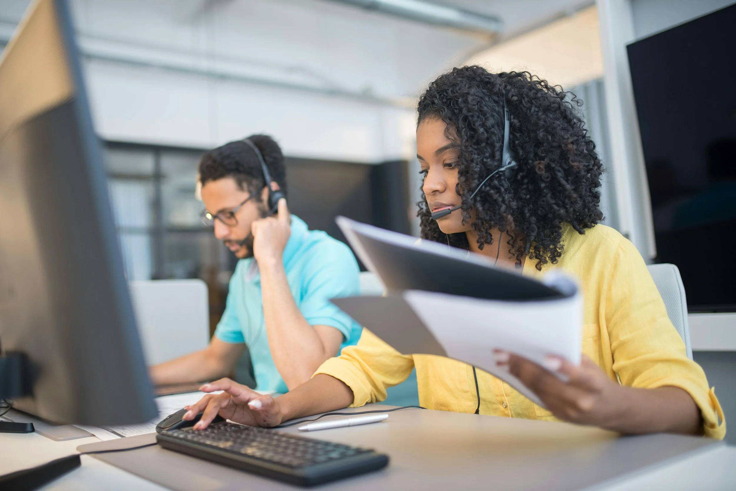 Two individuals wearing headsets working at their computers in an office setting, focused on their tasks.