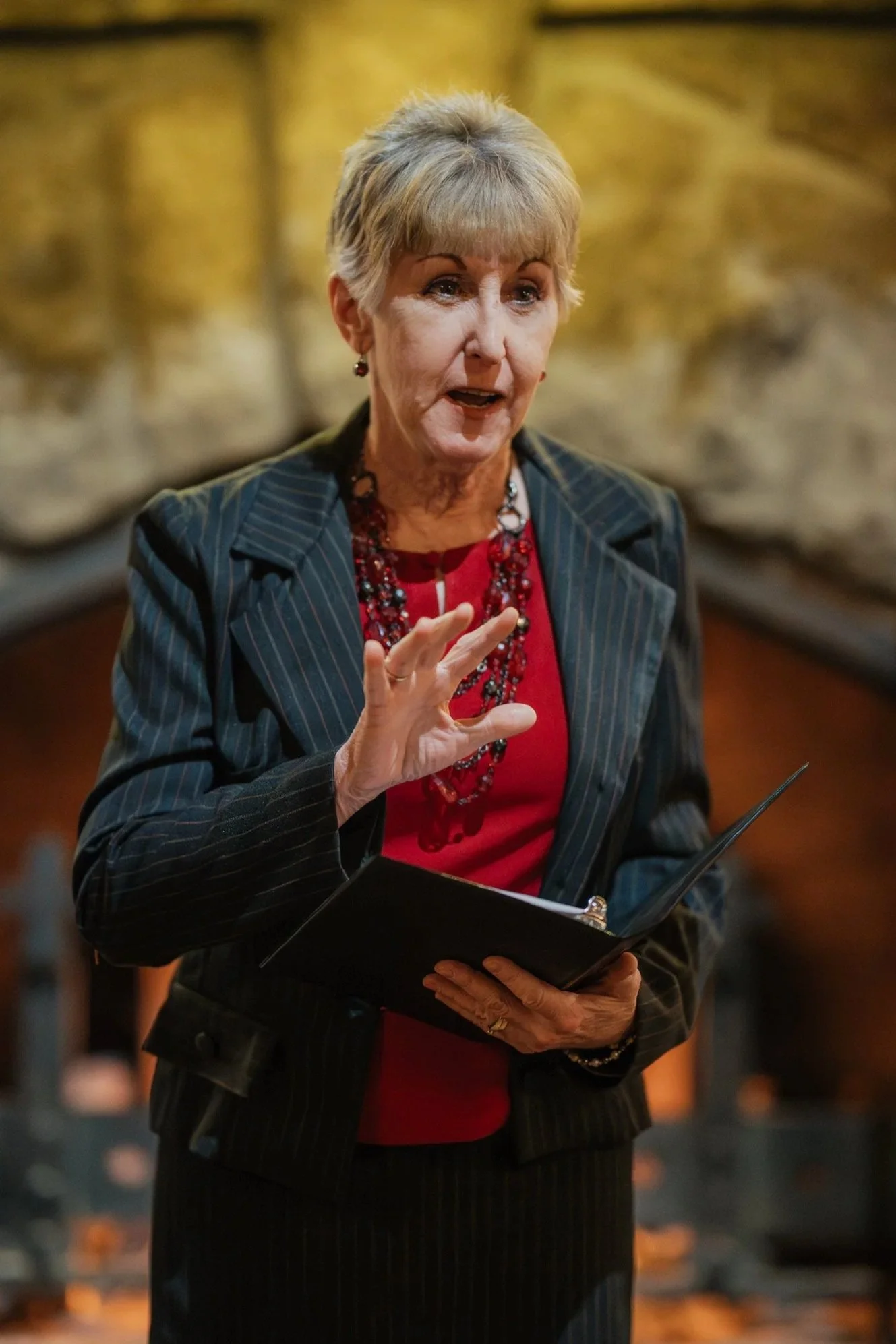 An older woman with short blonde hair, dressed in a pinstripe blazer and red top, speaking and gesturing with her hand while holding a folder.