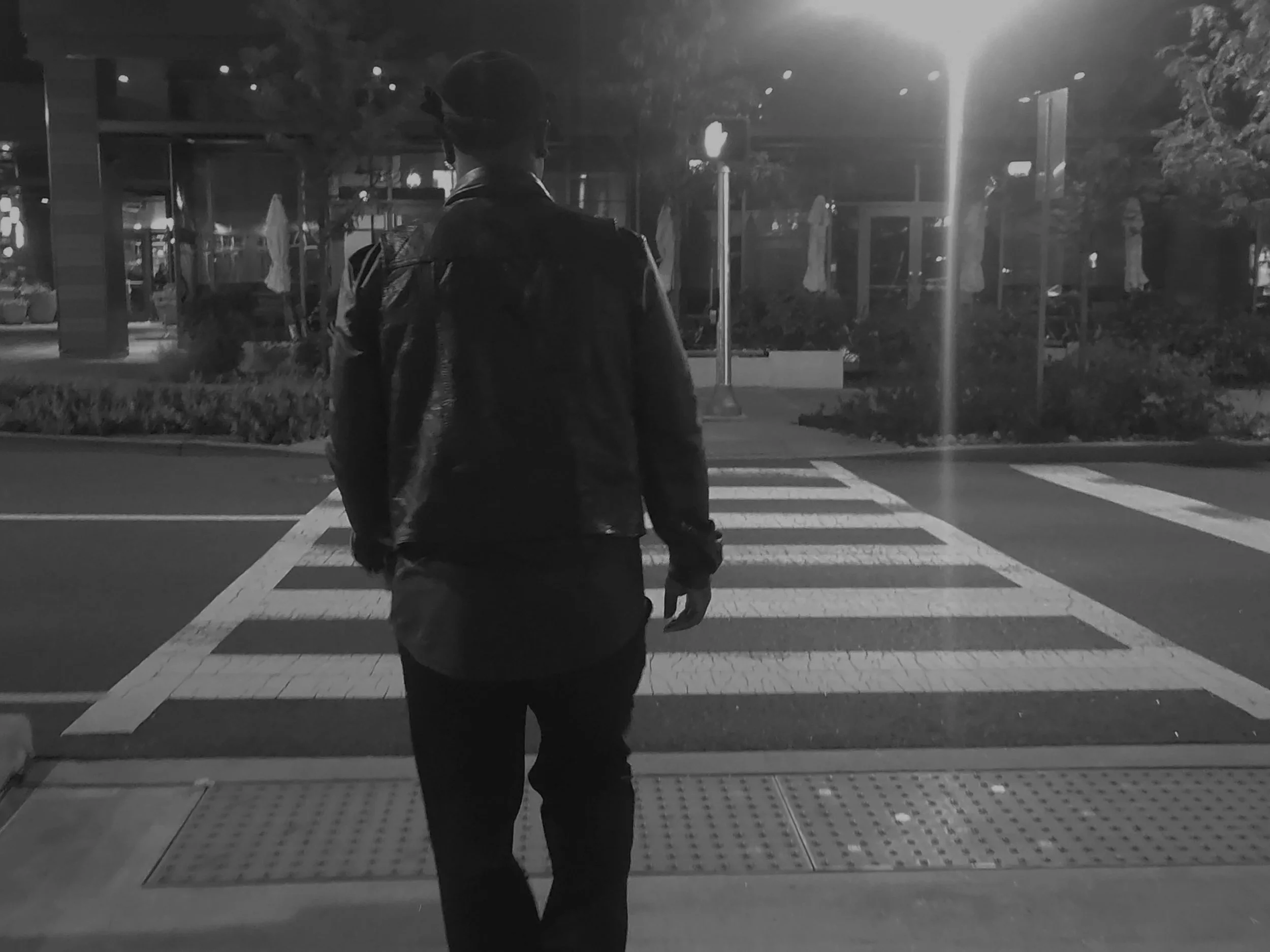 A person wearing a hooded jacket and carrying a backpack walking across a pedestrian crosswalk at night in an urban setting, illuminated by streetlights.