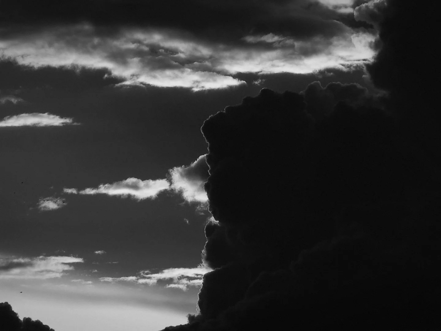 Black and white photo of a dramatic sky with large dark clouds, contrasted by smaller illuminated clouds, creating a striking silhouette effect.