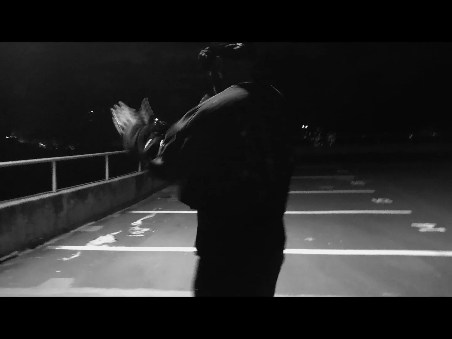 A person standing alone in a parking lot at night, wearing a jacket and cap, looking down at a fish they are holding.