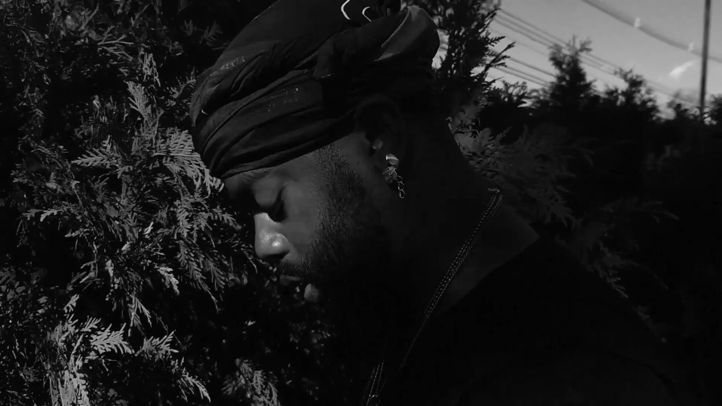 A man wearing a head wrap, earrings, and a dark jacket stands next to leafy foliage in black and white, with his face turned to the side and eyes closed.