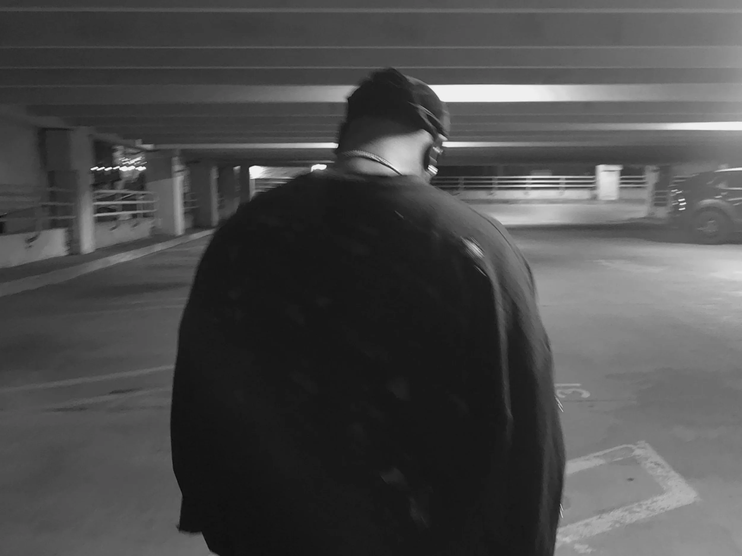 A person with short dark hair wearing a dark jacket and sunglasses, standing in an outdoor parking garage, facing away from the camera.