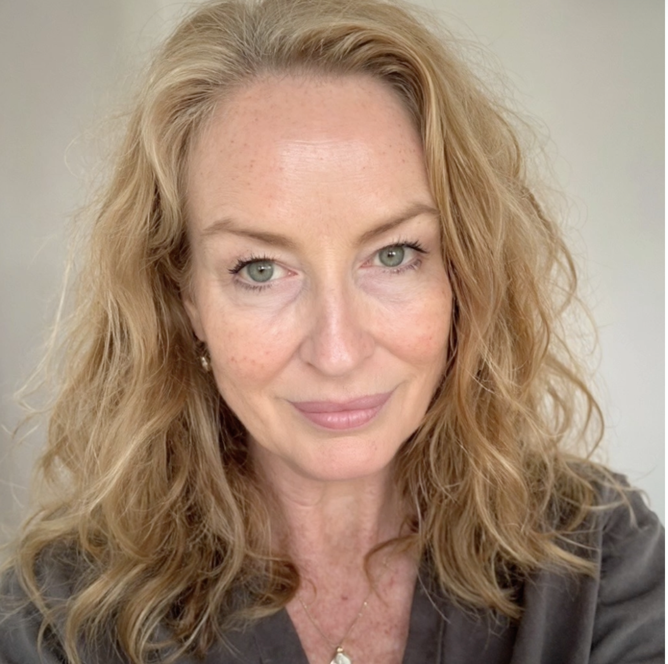 A woman with long, wavy red hair and green eyes, smiling softly, wearing a dark top and a pearl pendant necklace, standing against a plain light background.