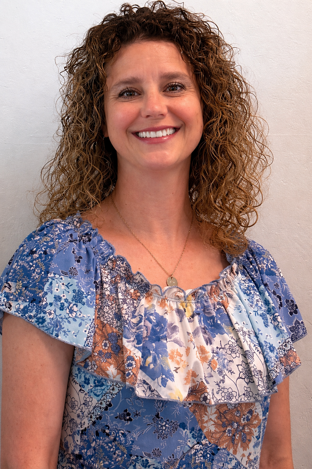 A woman with curly brown hair smiling, wearing a blue and floral patterned blouse and a gold necklace, standing against a plain white wall. Accountant, Bookkeeper, Bookkeeping, Accounting, Finances