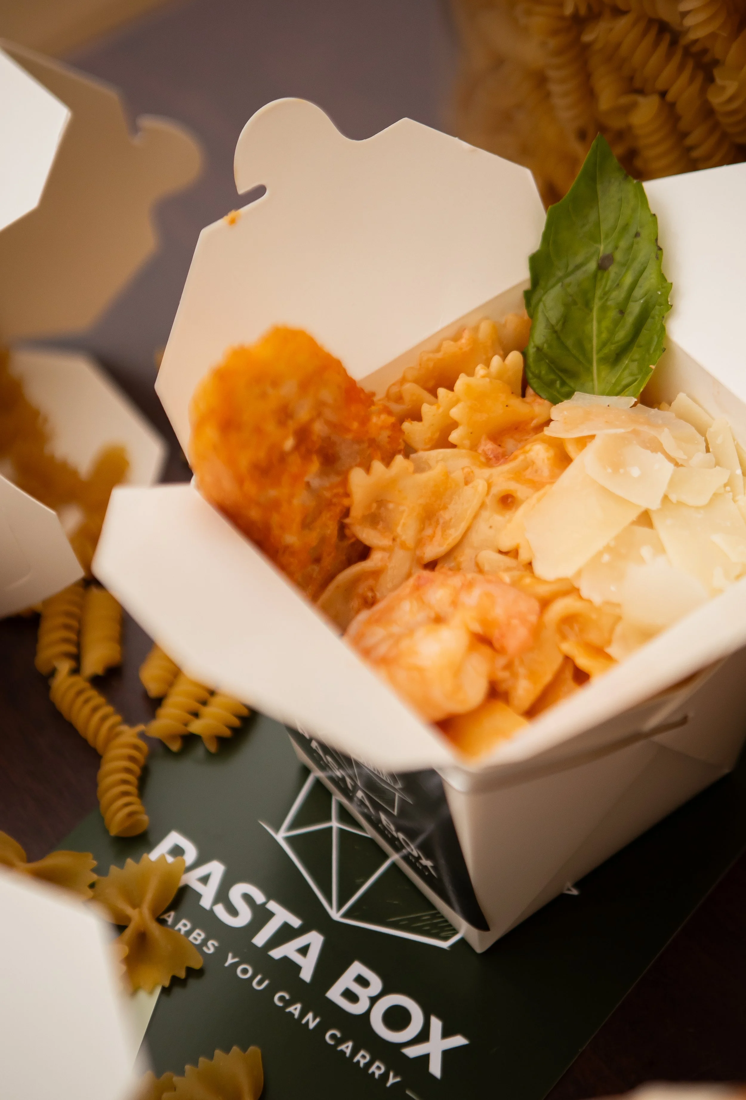 Close-up of a white takeout box filled with pasta, garnished with a basil leaf, and a piece of breaded chicken. There are uncooked fusilli pasta noodles around the box and a black paper bag beneath it with white text reading 'PASTA BOX' and a slogan.
