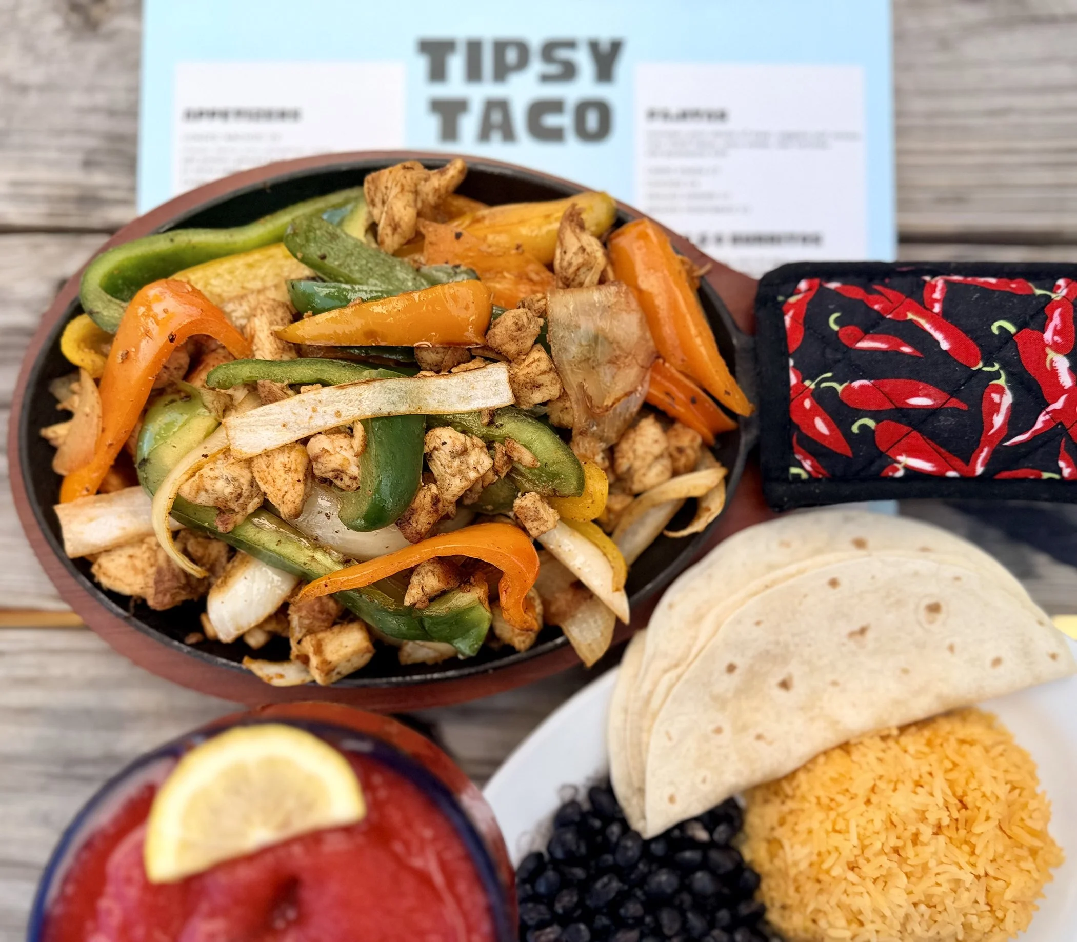 A bowl of stir-fried chicken and colorful bell peppers on a wooden table with a Tipsy Taco menu in the background.