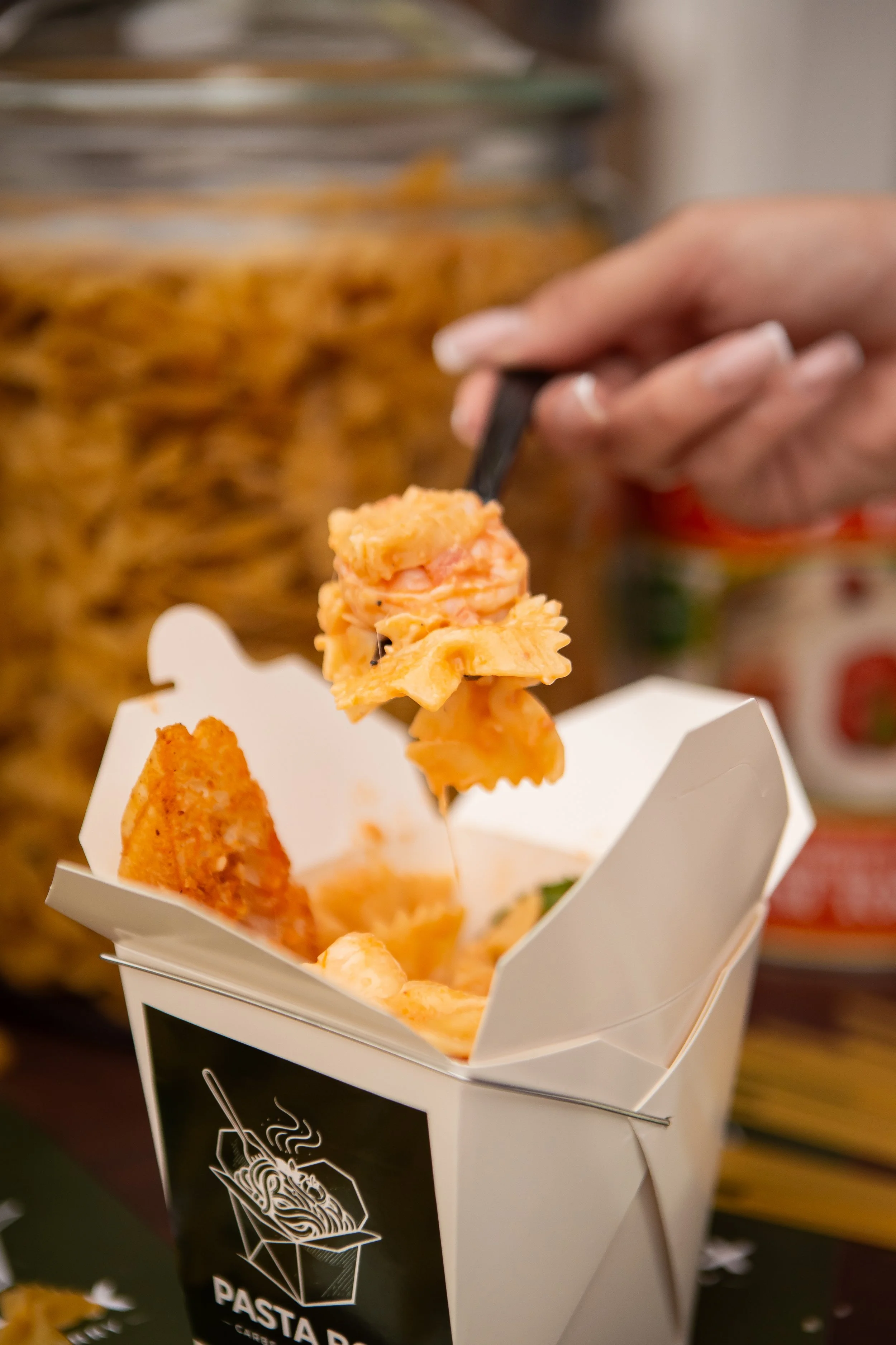 A hand holding black chopsticks picking up pasta with creamy orange sauce from a white takeout container.