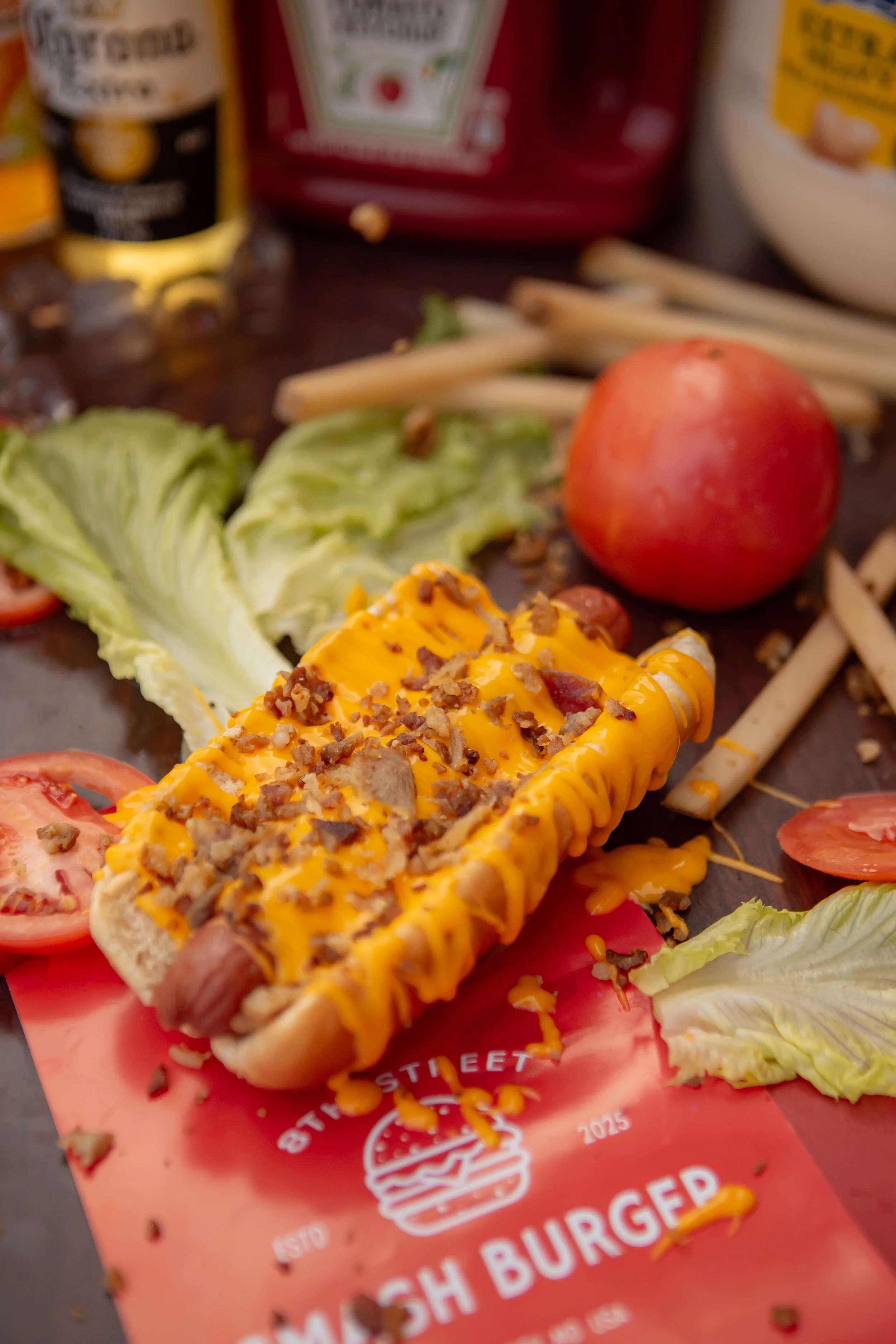 Close-up of a cheeseburger with lettuce, tomato, and cheese topped with chopped nuts, placed on a red burger wrapper with white text.