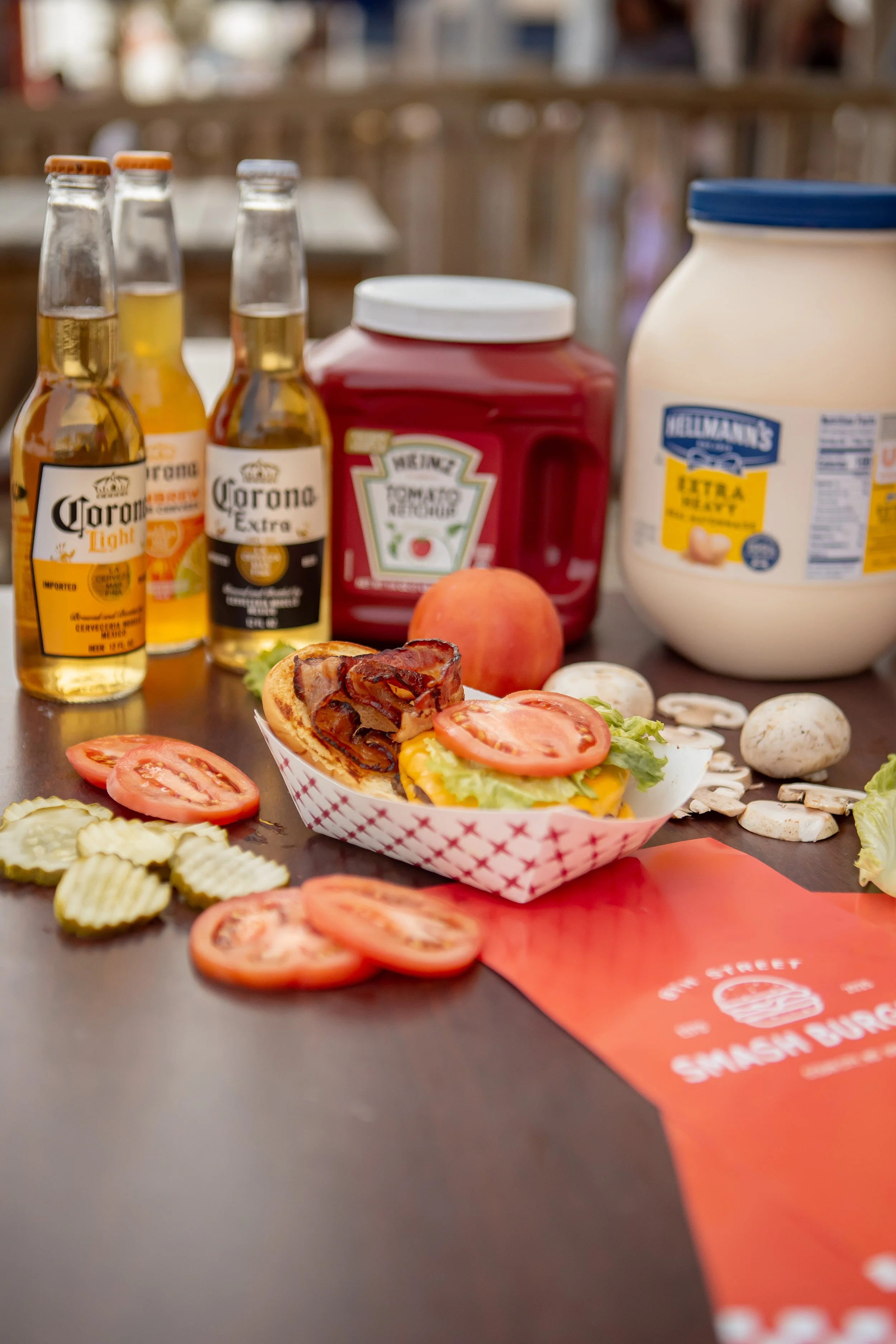 A table with bottles of Corona beer, a container of tomato ketchup, a jar of mayonnaise, sliced tomatoes, lettuce, mushrooms, slices of cucumber, slices of ham, pizza slices, and a red paper with white text.