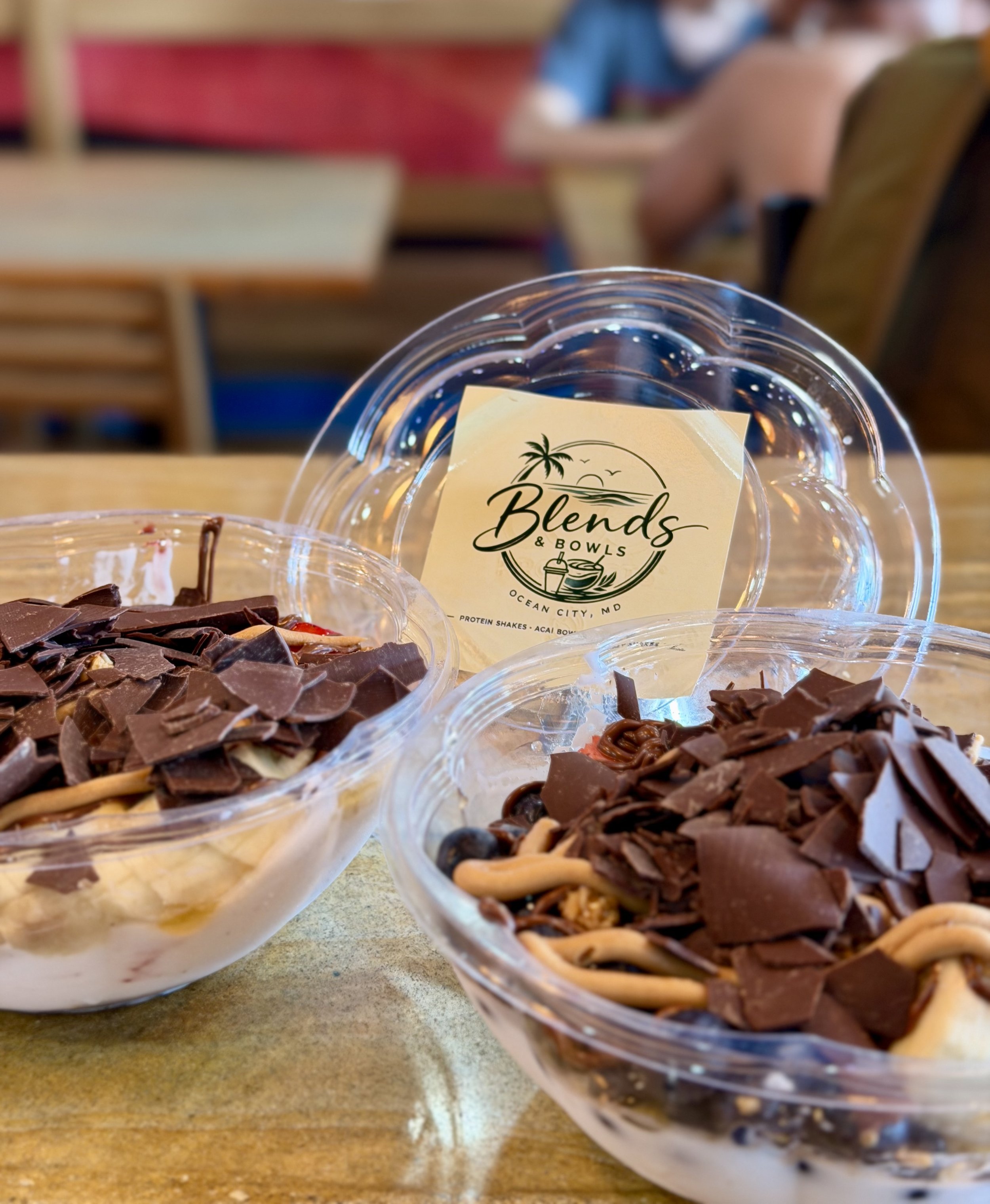 Close-up of chocolate desserts with chocolate shavings in clear plastic bowls on a wooden table, with a blurred restaurant setting in the background.