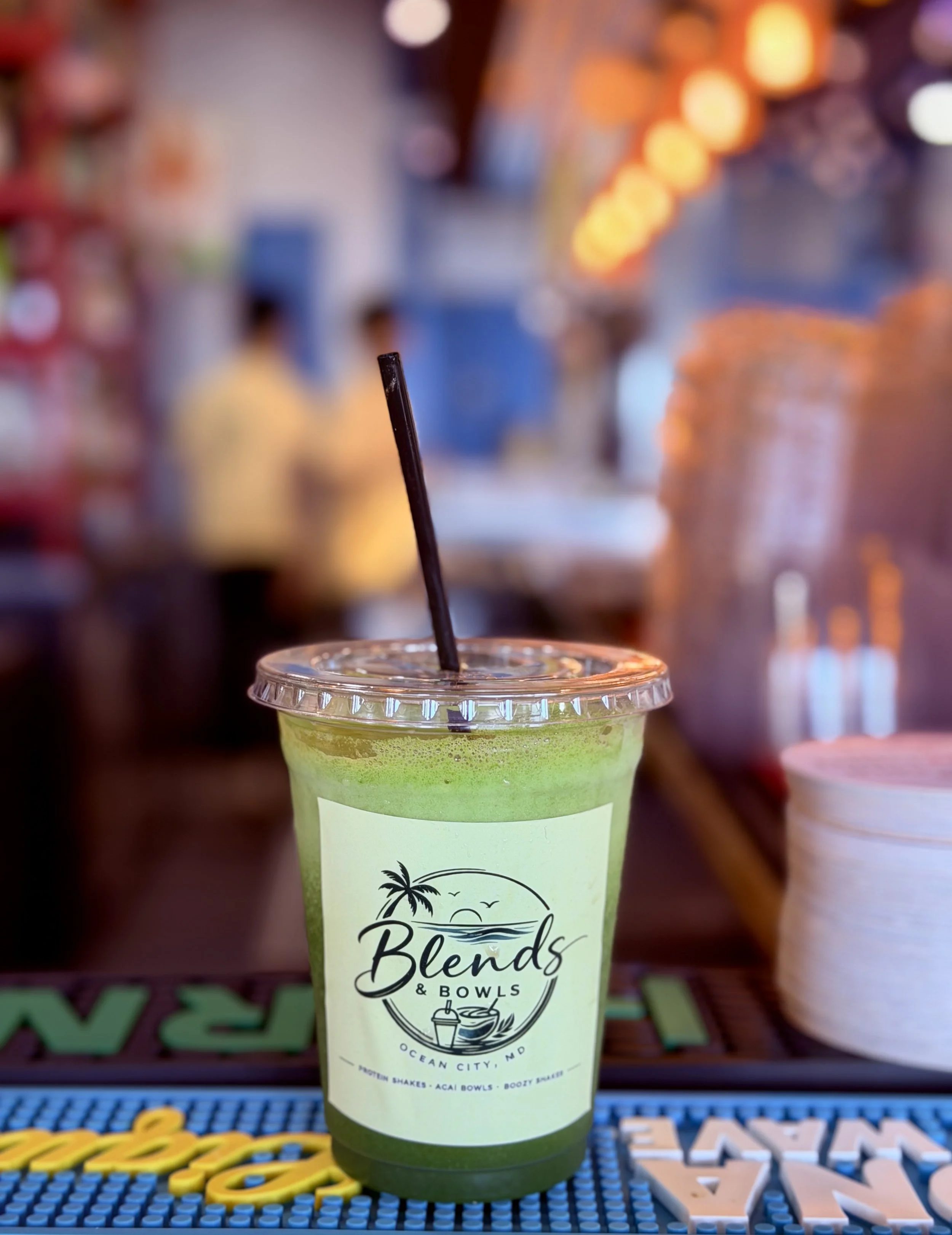 A green smoothie in a clear plastic cup with a black straw, branded with a logo for 'Beaches & Bowls' that includes a palm tree and an ocean scene, placed on a colorful surface with blurred lights and decor in the background.