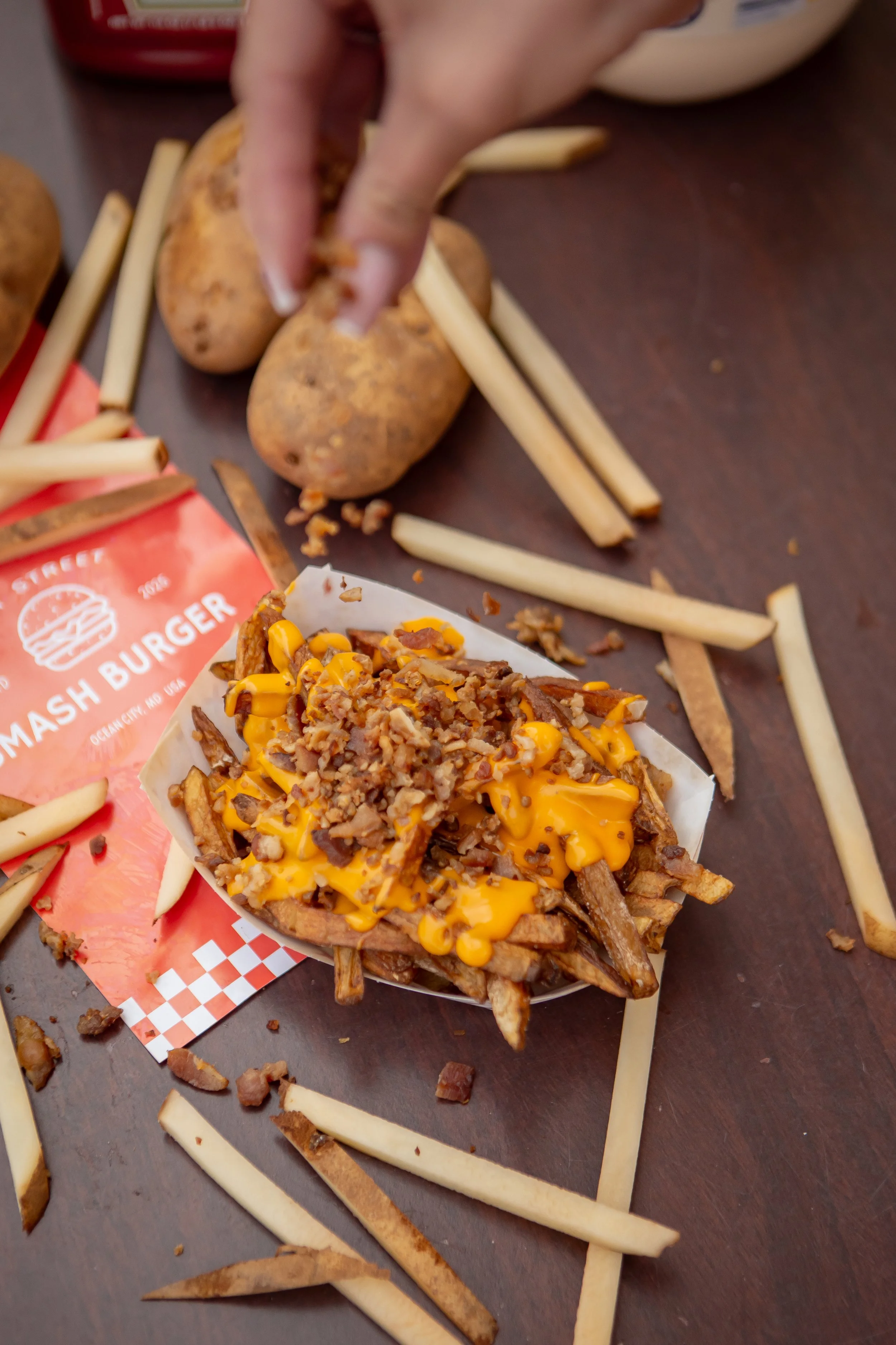 A tray of loaded French fries topped with shredded cheese, bacon bits, and shredded meat. In the background, there are potatoes, fries, and some sauce containers on a dark surface.