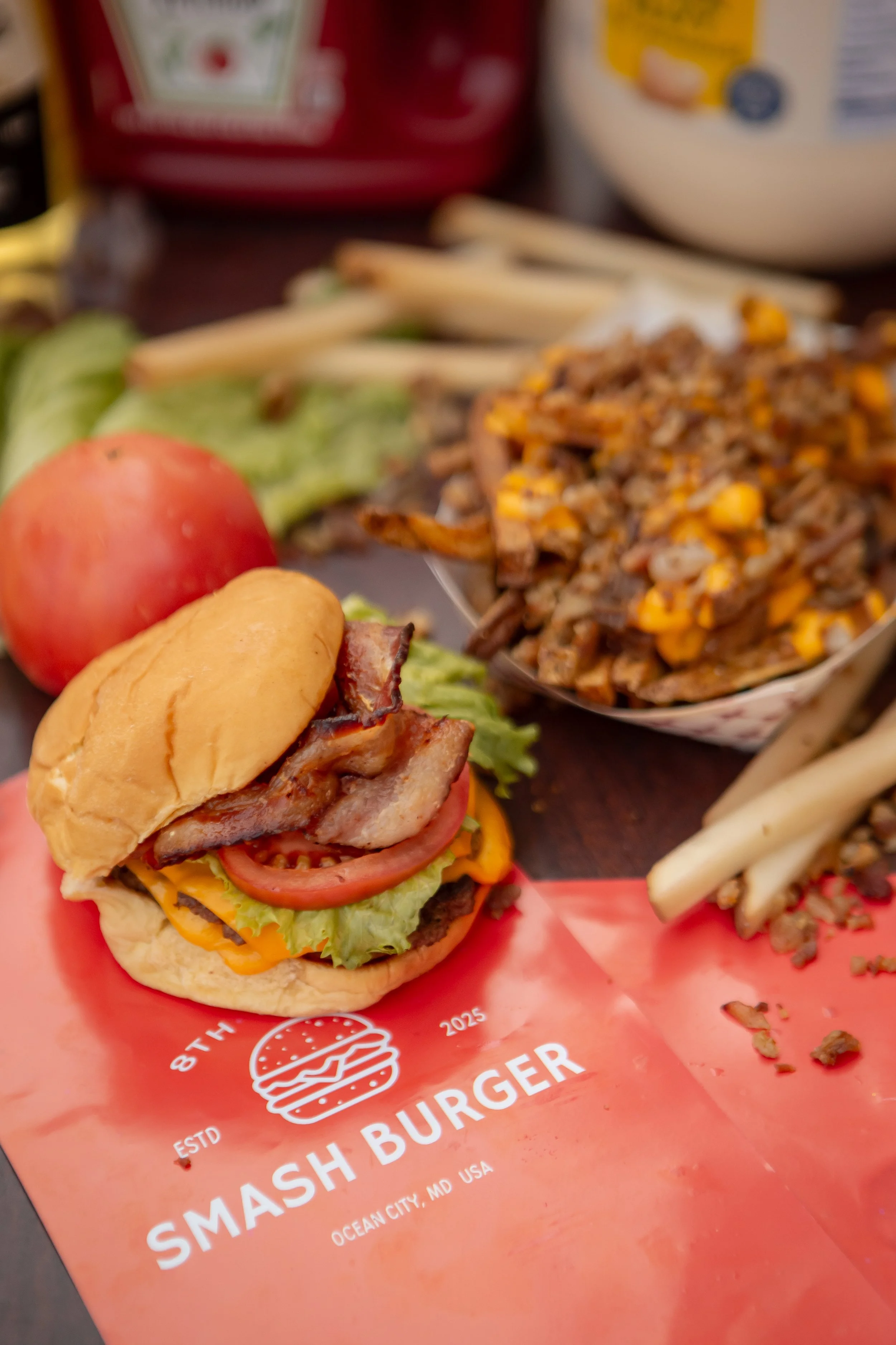 A cheeseburger with bacon, lettuce, tomato, and cheese on a bun, placed on a red paper wrapper with white text and a burger logo, along with a side of French fries, rice, and a bowl of rice and beans, with condiments in the background.