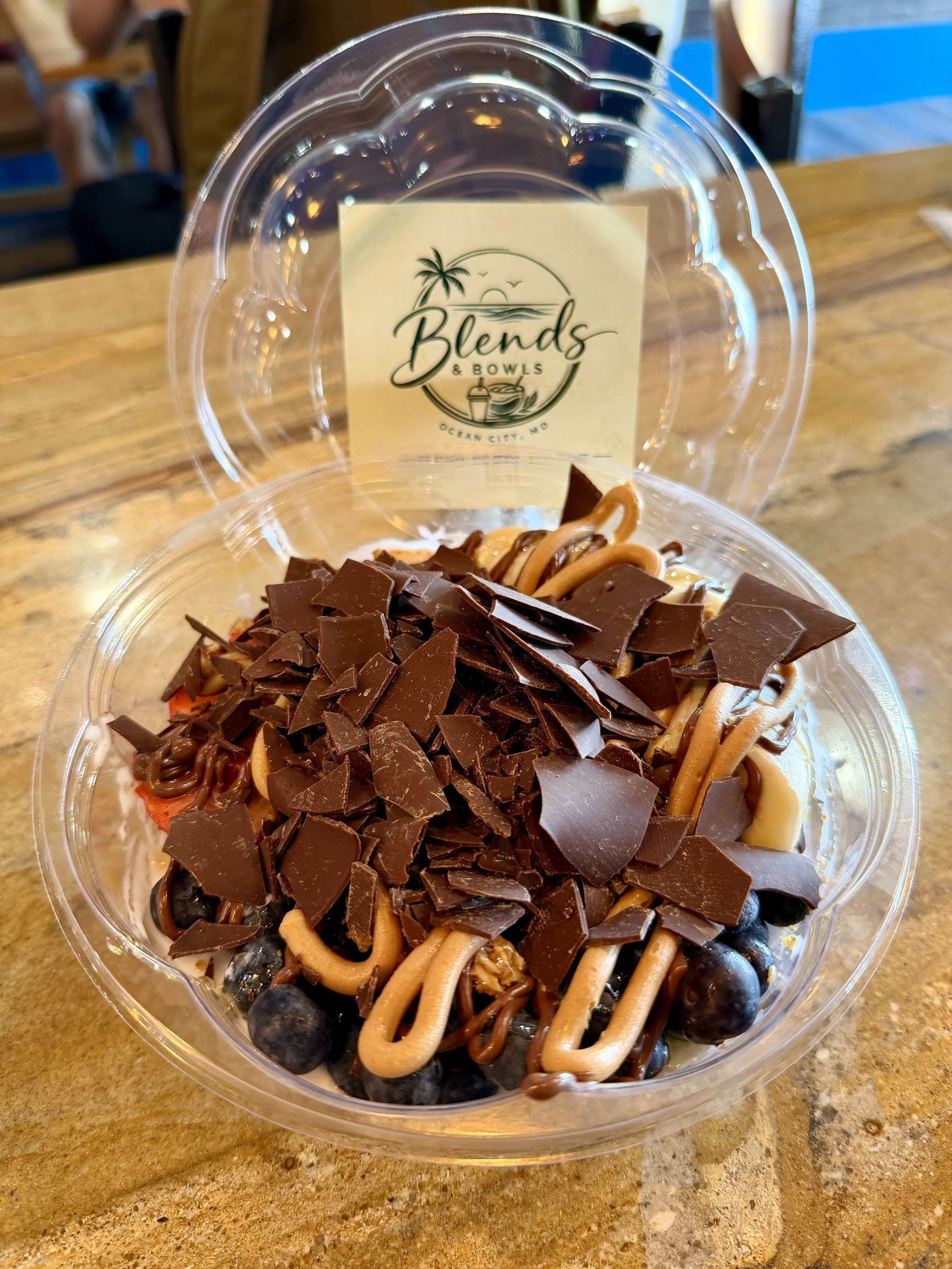 A clear plastic takeout container filled with a dessert topped with chocolate shavings, sitting on a wooden table. In the background, a sticker on the lid reads 'Blends & Bowls, Ocean City, MD' with a design of a palm tree, sunset, and ocean waves.