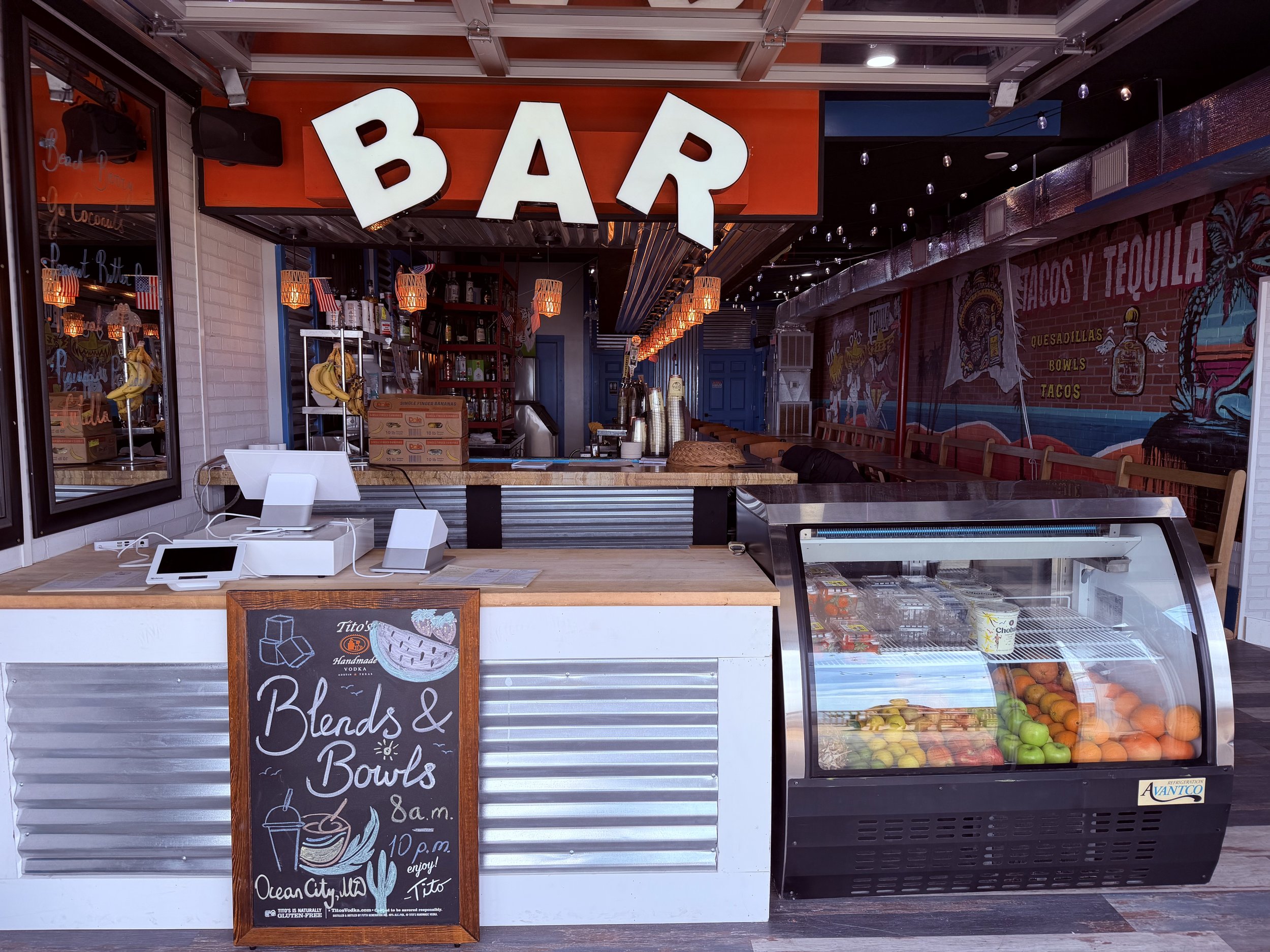 The image shows a food and drink stand with a bright orange sign that reads 'BAR' in white capital letters. Inside, there are bottles on shelves, hanging lanterns, and a menu board with handwritten text promoting blended drinks and bowls. A glass dis