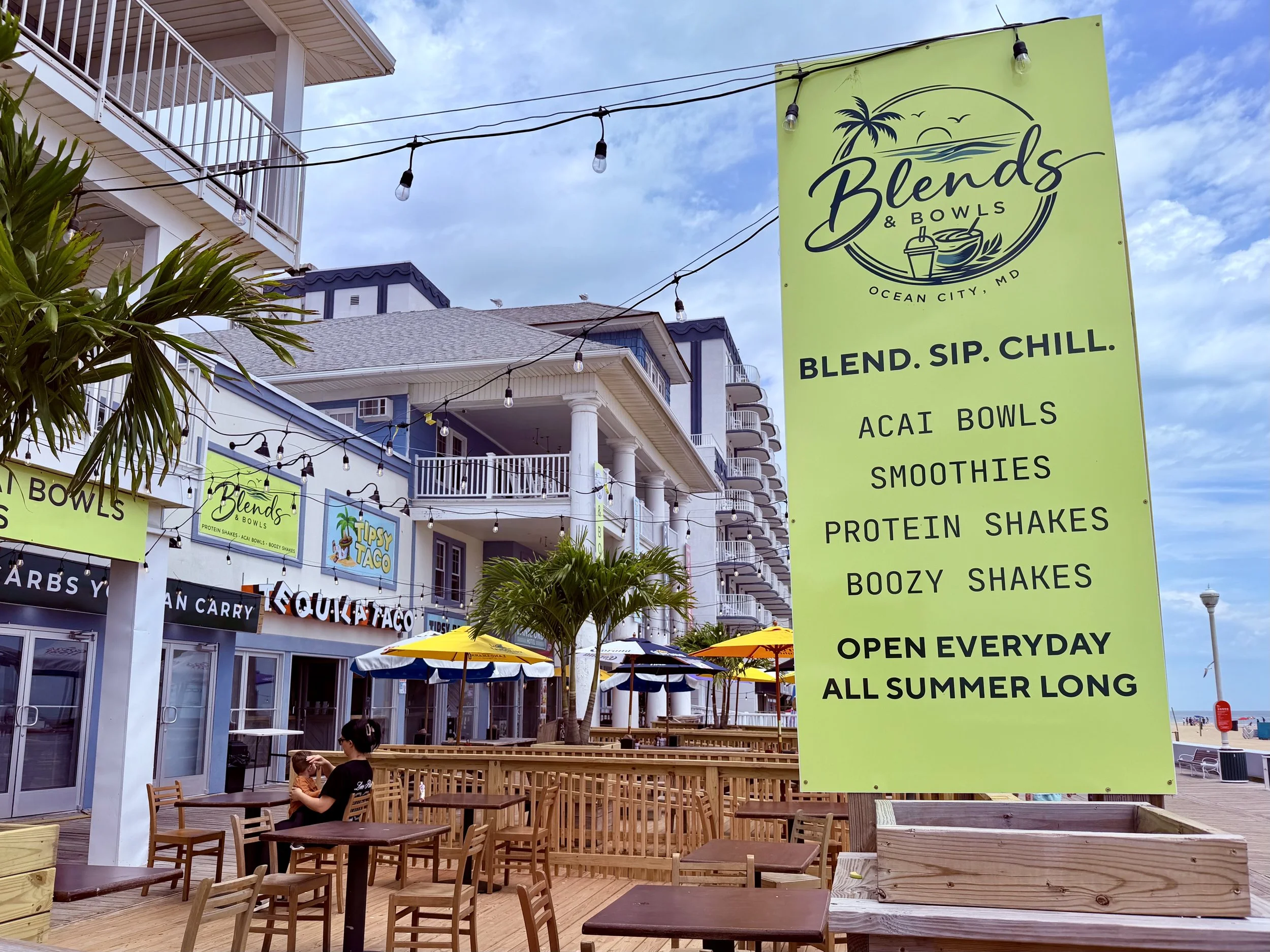 Bright green sign outside a restaurant called Blends & Bowls in Ocean City, Maryland, advertising their menu including acai bowls, smoothies, protein shakes, buzy shakes, and indicating they are open every day of summer.