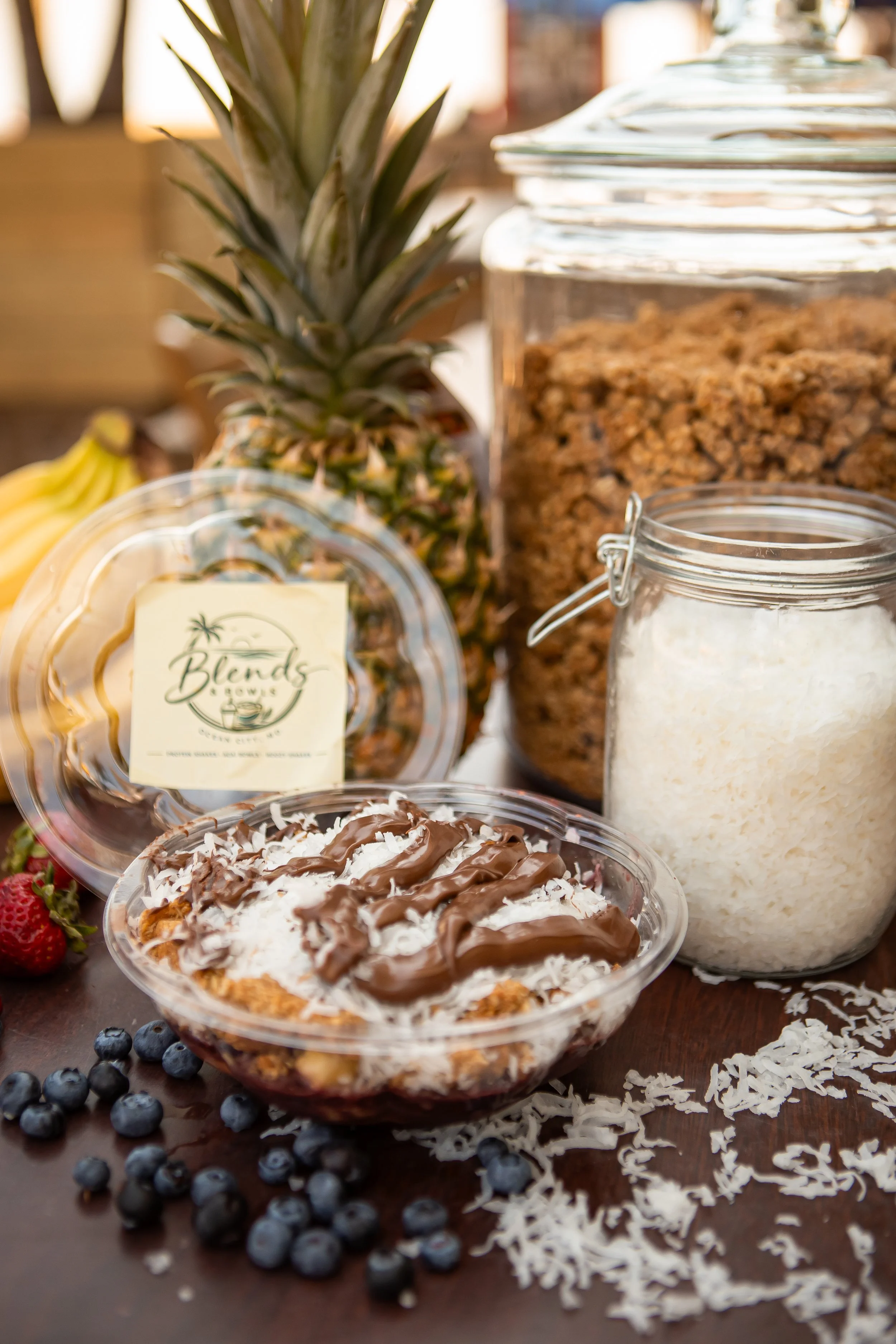A bowl of coconut and chocolate dessert with shredded coconut on top. Fresh blueberries scattered on the table, a pineapple and banana in the background, and glass jars filled with granola and cereal nearby.