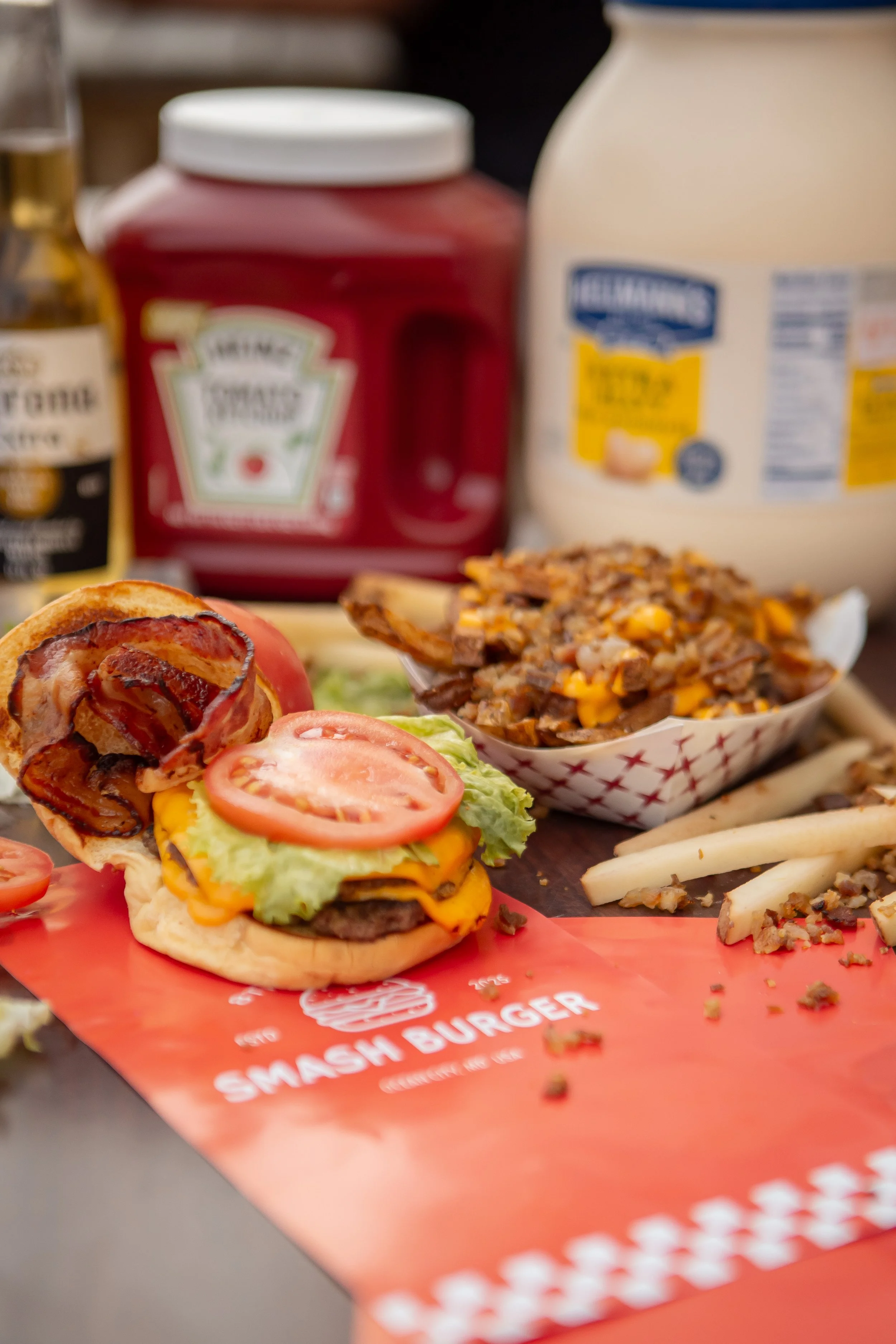 Close-up view of a cheeseburger with bacon, tomato, lettuce, and cheese on a bun, placed on a red plate with a menu and condiments in the background.