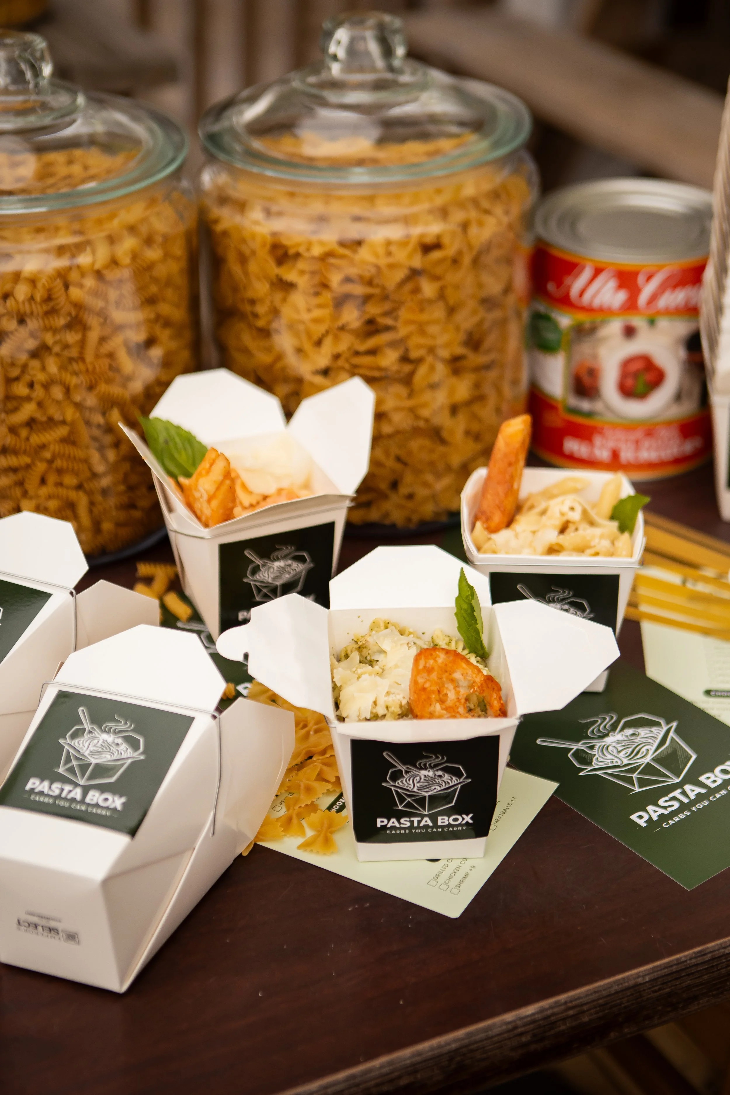 Small white takeout boxes with black labels reading "PASTA BOX" and images of pasta, surrounded by large glass jars filled with different types of dry pasta on a wooden table.