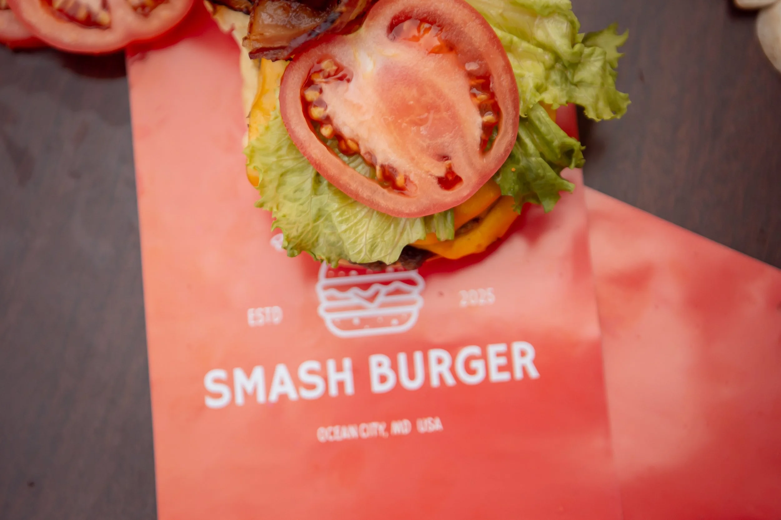 Close-up of a burger with tomato, lettuce, bacon, and cheese on a bun, placed on a red surface with branding text in the background.