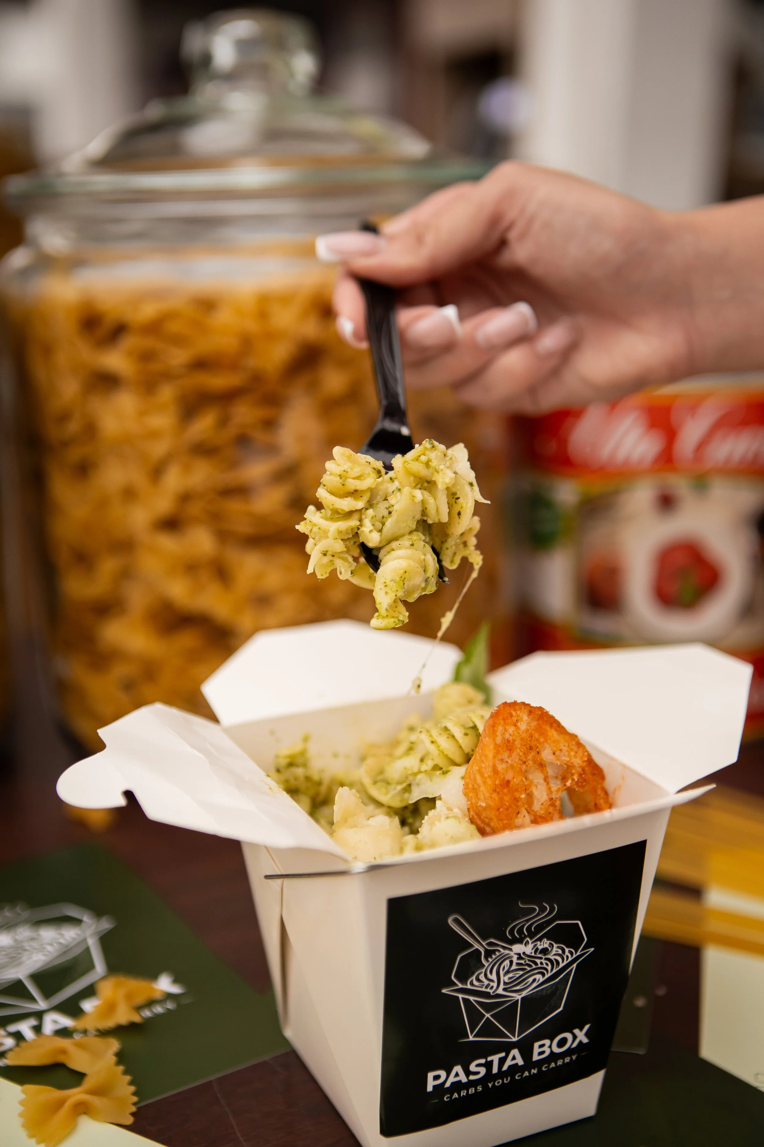 A container of pasta from Pasta Box, with a hand holding a forkful of pasta topped with herbs over a takeout box. In the background, there is a jar of pasta and other items.