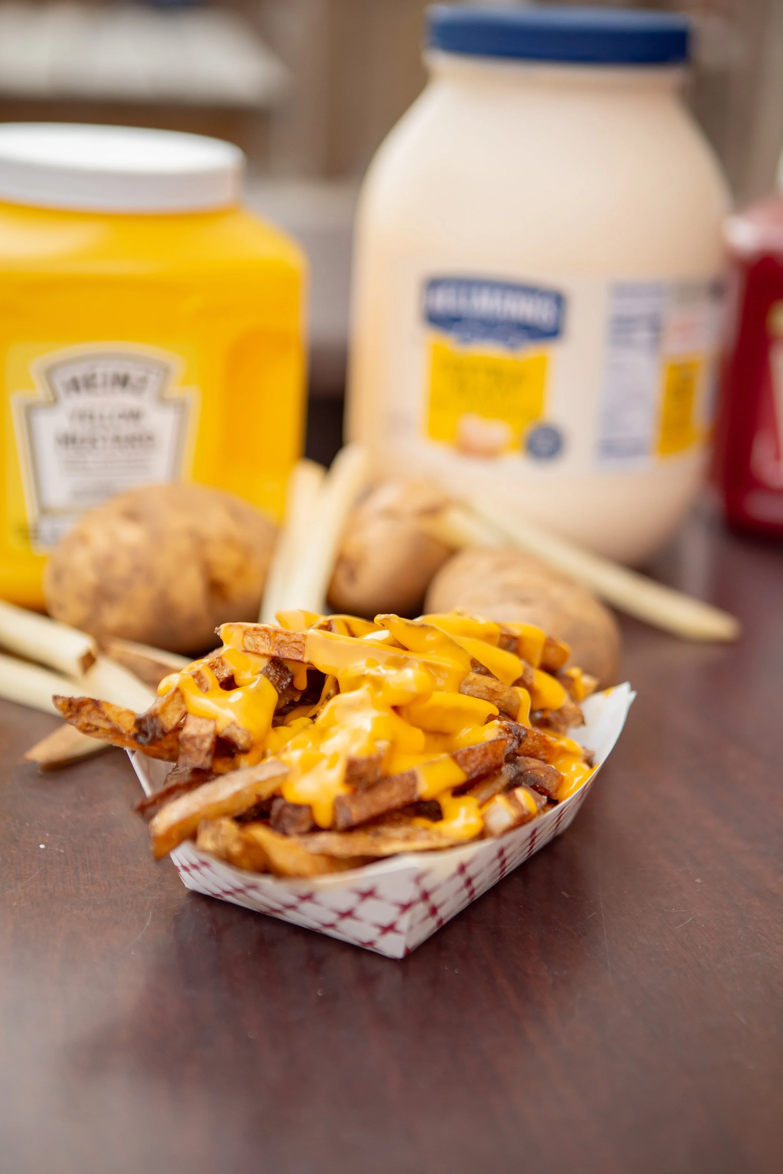 A serving of french fries topped with melted cheddar cheese in a paper basket, with baked potatoes, bottles of mayonnaise and mustard in the background.