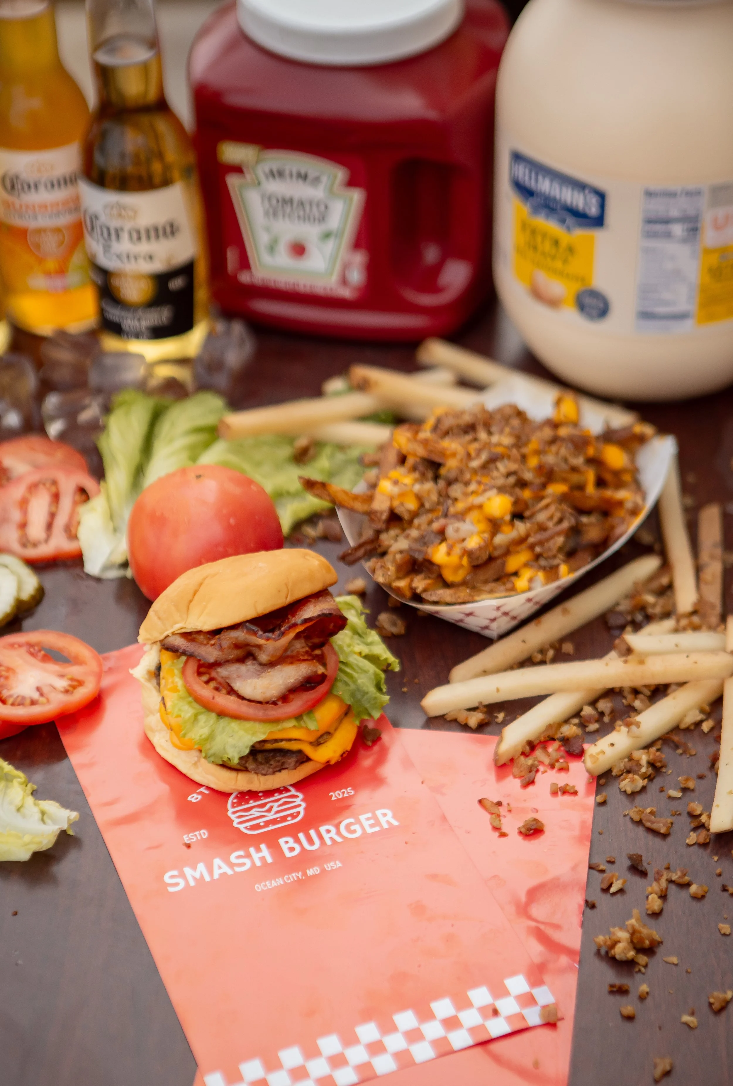 A table with a hamburger slider, lettuce, tomato, and onion slices, topped with bacon, and a serving of chili with corn, alongside french fries and scattered ground meat. There are condiment bottles, a red ketchup dispenser, a white mayonnaise jar, a