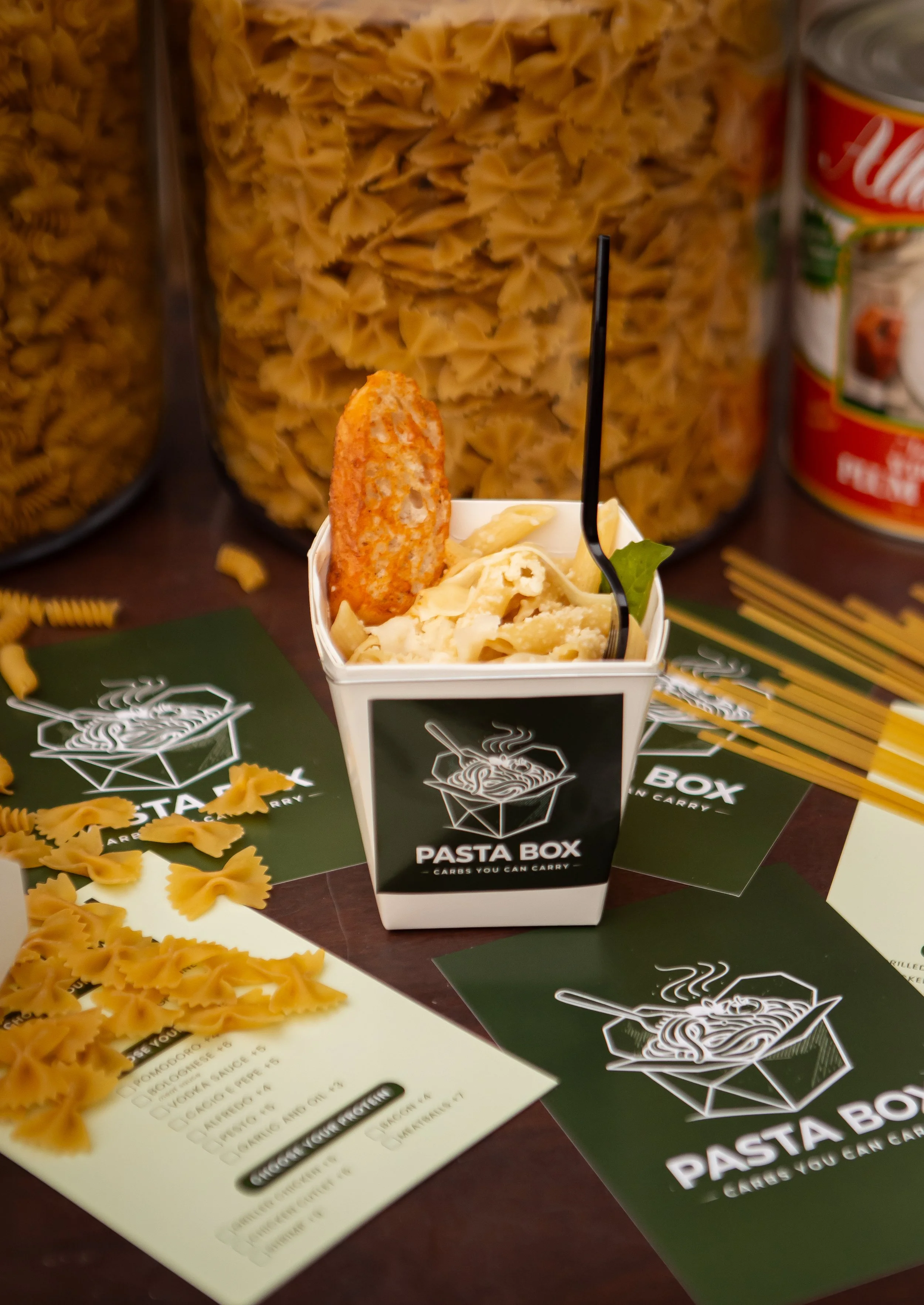 A container of pasta with a breaded chicken strip, surrounded by packets and jars of pasta and sauce, on a table with black and green informational cards that have white text and graphics.