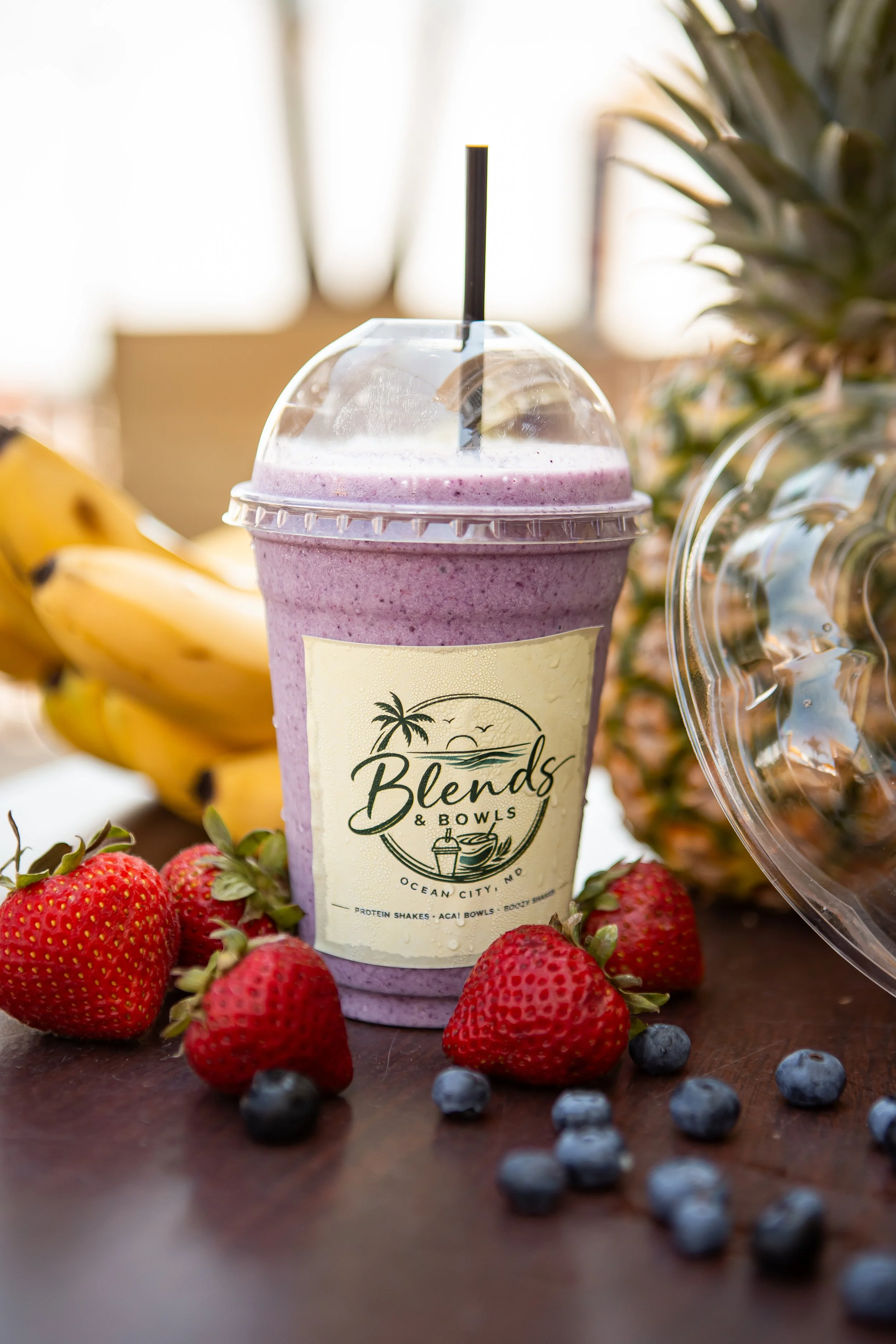 A purple smoothie in a clear plastic cup with a black straw, surrounded by strawberries and blueberries, with pineapple and banana in the background, on a wooden surface.