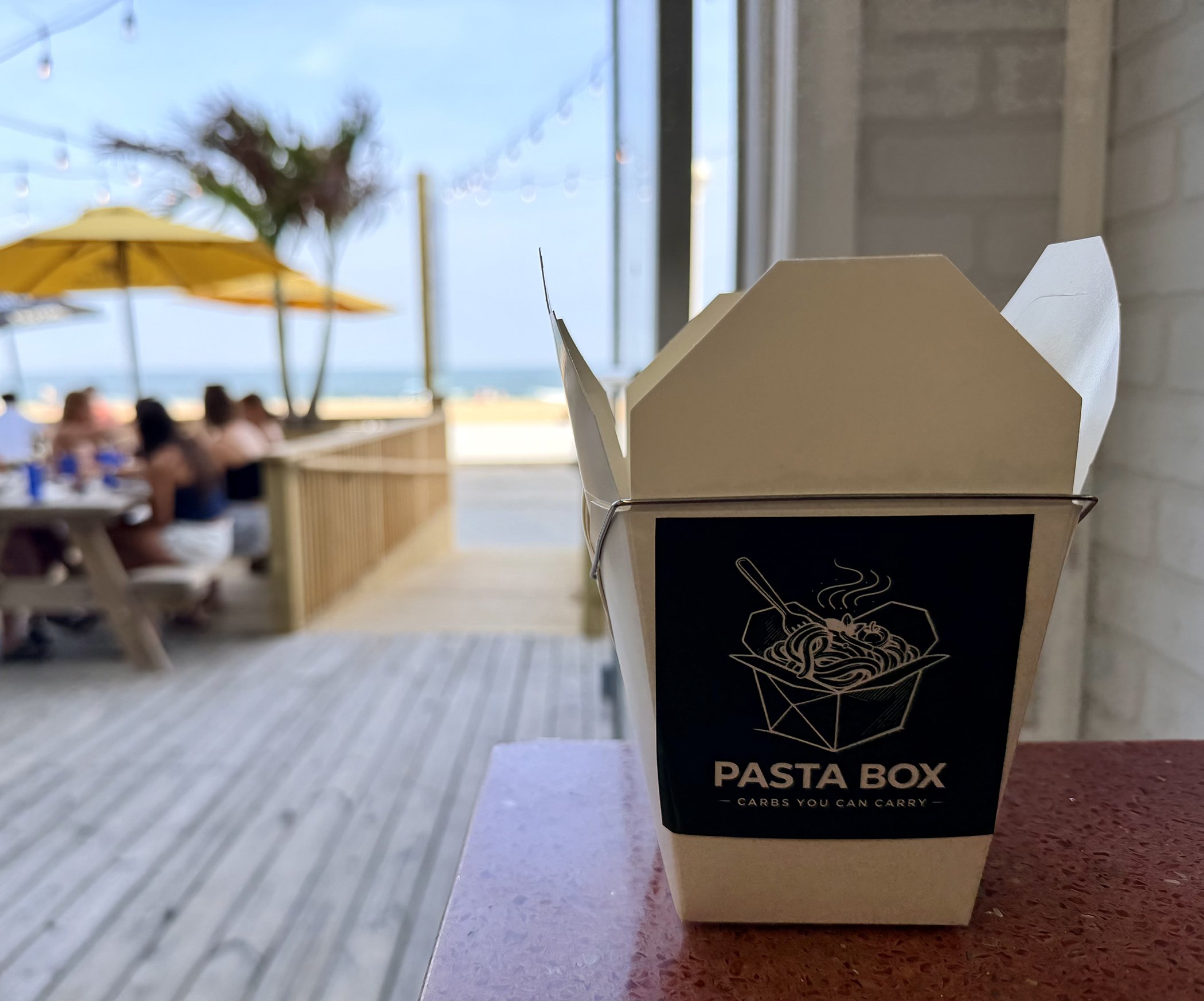 A takeout pasta container on a black pasta box with a design of a spider web and some text, positioned on a table in a restaurant or cafe with a beach view, yellow umbrellas, and people sitting in the background.