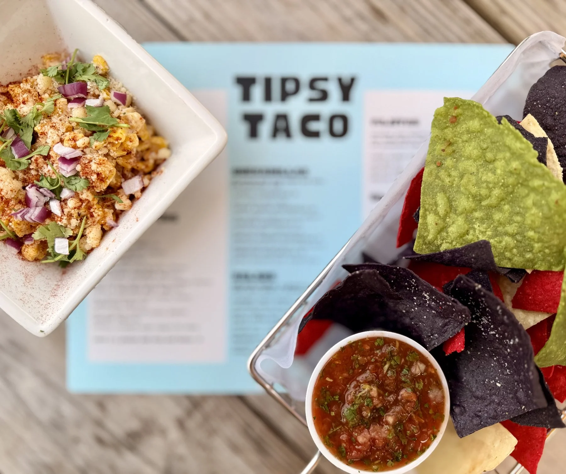 Close-up of a tray of nachos topped with green and black tortilla chips, with sides of salsa and a bowl of chopped vegetable salsa. In the background, a greeting card with the words 'TIPSY TACOS' is visible.