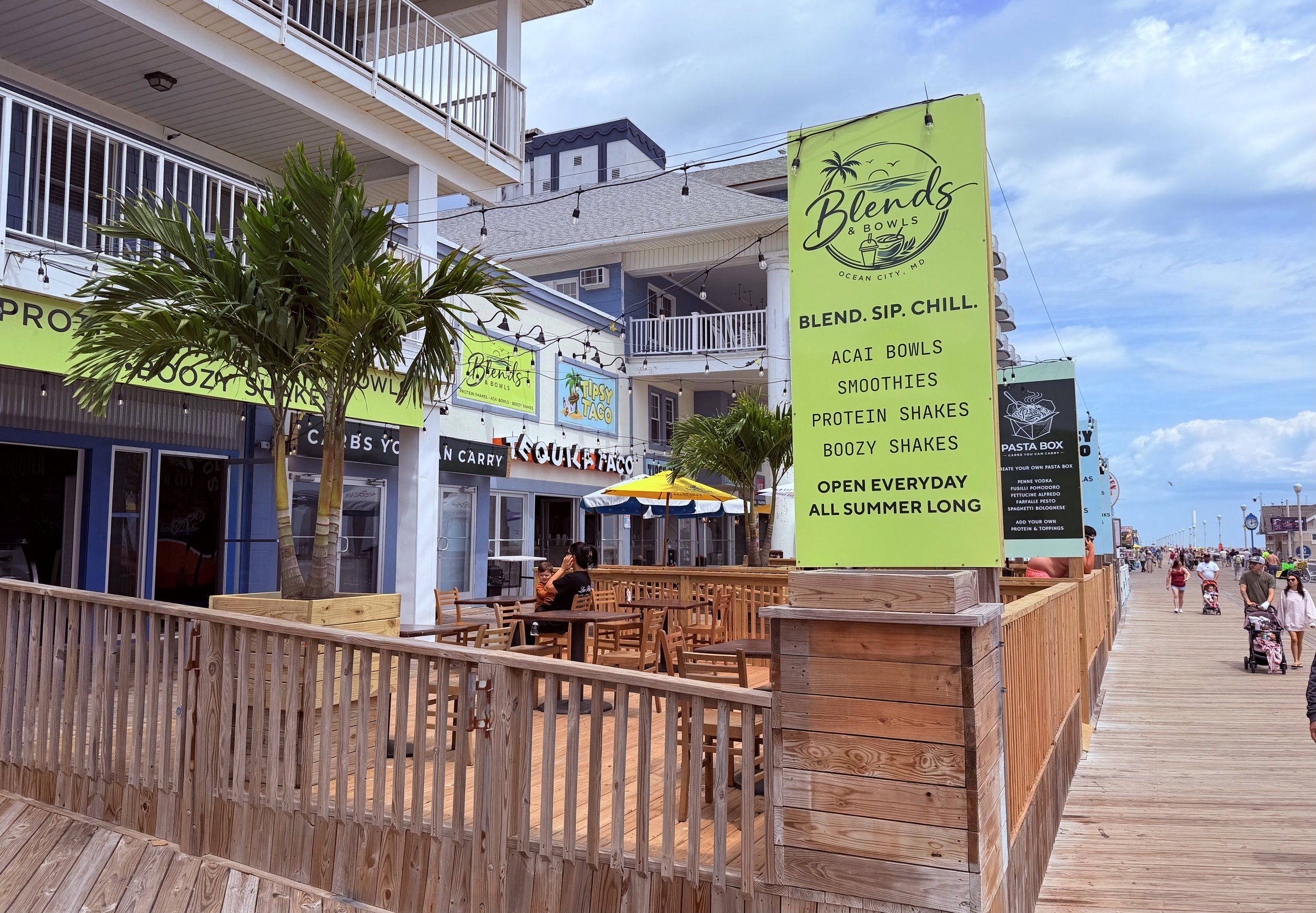 Outdoor shopping and dining area with a green banner advertising Blends & Bowls, a smoothie shop in Ocean City, MD, featuring smoothie, acai bowls, protein, and boozy shakes, with people walking on a boardwalk and a cloudy sky.