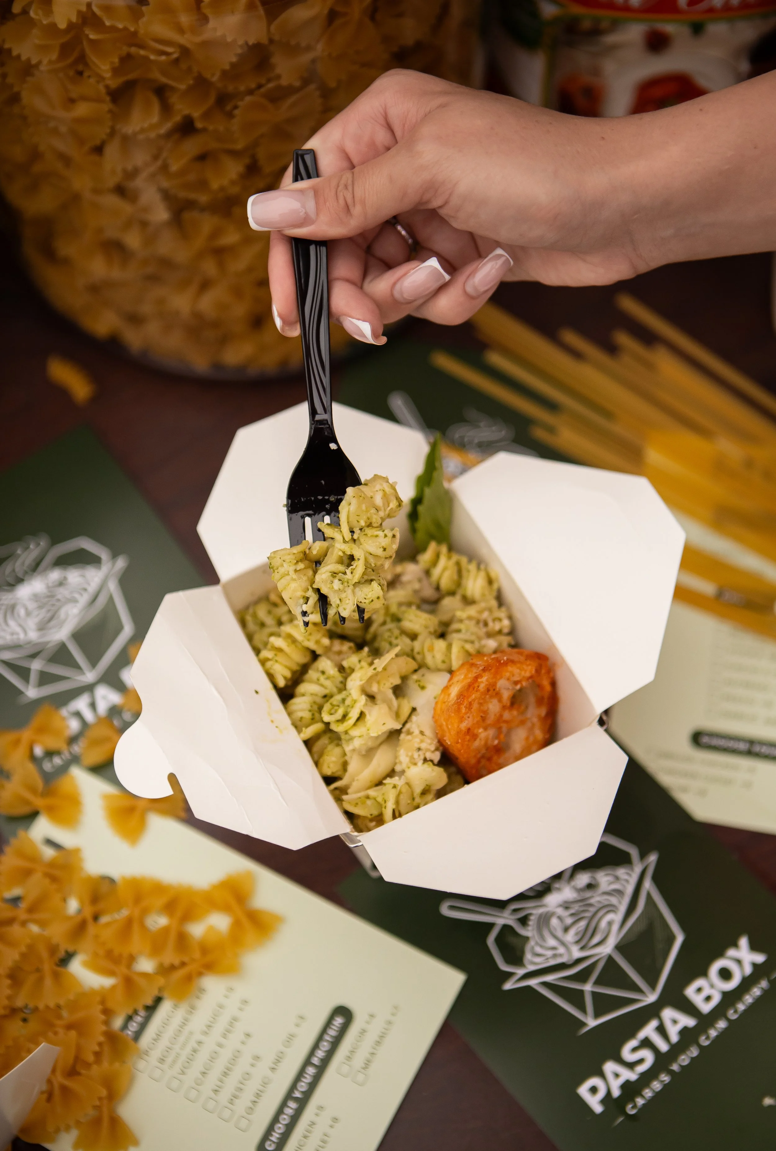 Hand holding a fork grabbing pasta from a takeout box with pasta salad and a meatball inside, in front of pasta packaging and a menu in the background.