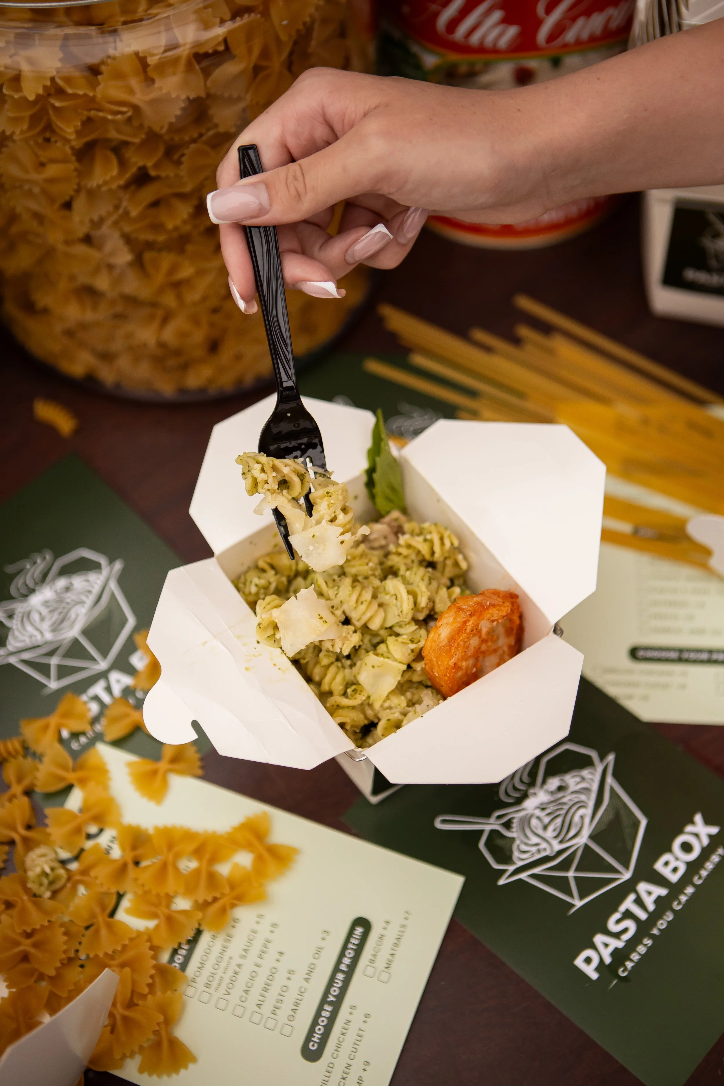 A hand holding a black fork with a piece of pasta and a piece of fried food in a white takeout container with a green leaf, placed on a table with pasta and a menu from Pasta Box.